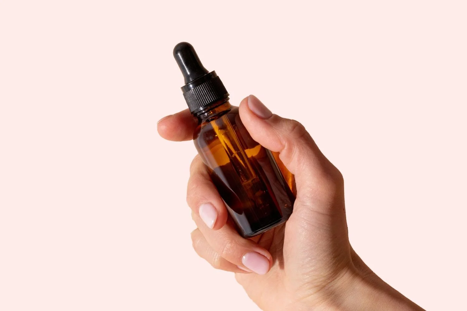 A hand holding a small amber glass dropper bottle with a black cap and rubber bulb, against a light pink background.
