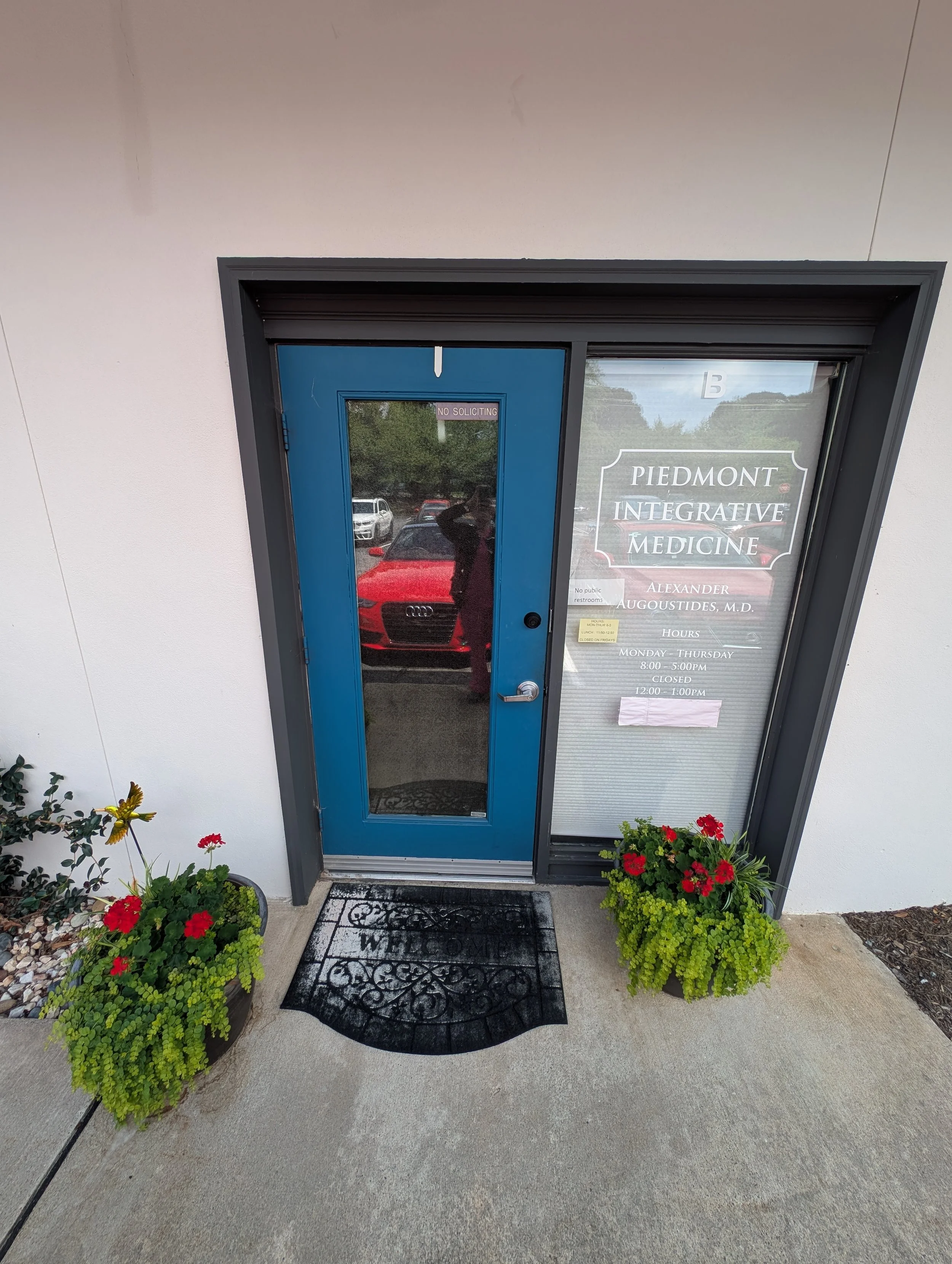 The entrance door of Piedmont Integrative Medicine with a sign indicating hours and the practitioner's name, Alexander Augoustides, M.D., flanked by potted plants and a mat with a welcoming message.