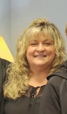A smiling woman with curly blonde hair, wearing a black top, standing indoors.