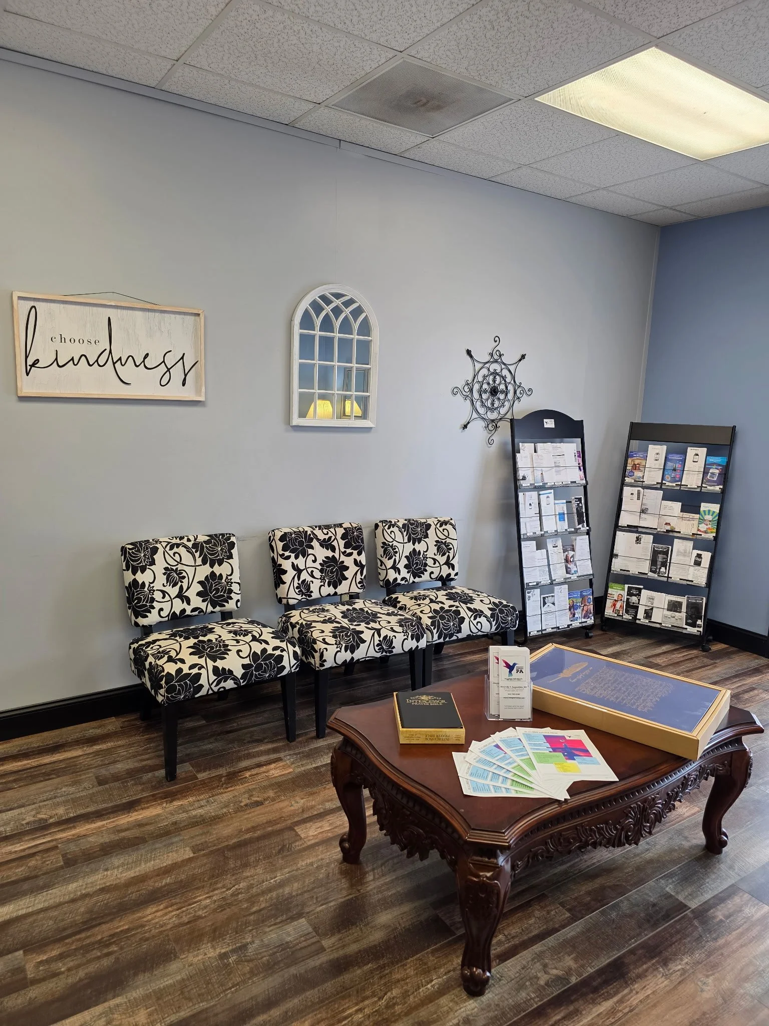 An office waiting area with three black and white floral patterned chairs pushed against a light gray wall. A wooden table in front holds reading materials and brochures. To the right, there are two black display racks with pamphlets. The wall has a decorative wall piece and a sign that says 'choose kindness'. The ceiling has white acoustic tiles with a fluorescent light.