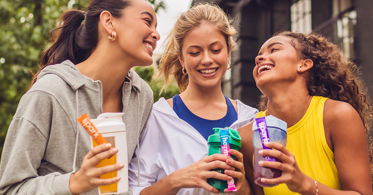 Three young women smiling and laughing outdoors, holding water bottles and energy drinks