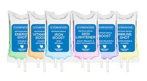 A row of six infusion bags with blue labels, each labeled with different health supplements including Energy Shot, Vitamin Boost, Iron Boost, Skin Lightener, Immune Shot, and another supplement.