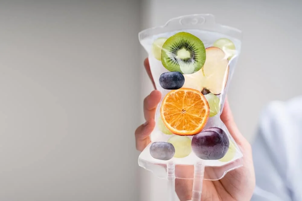 A clear plastic bag filled with assorted sliced fruits, including kiwi, orange, blueberries, and green grapes, being held up by a person.