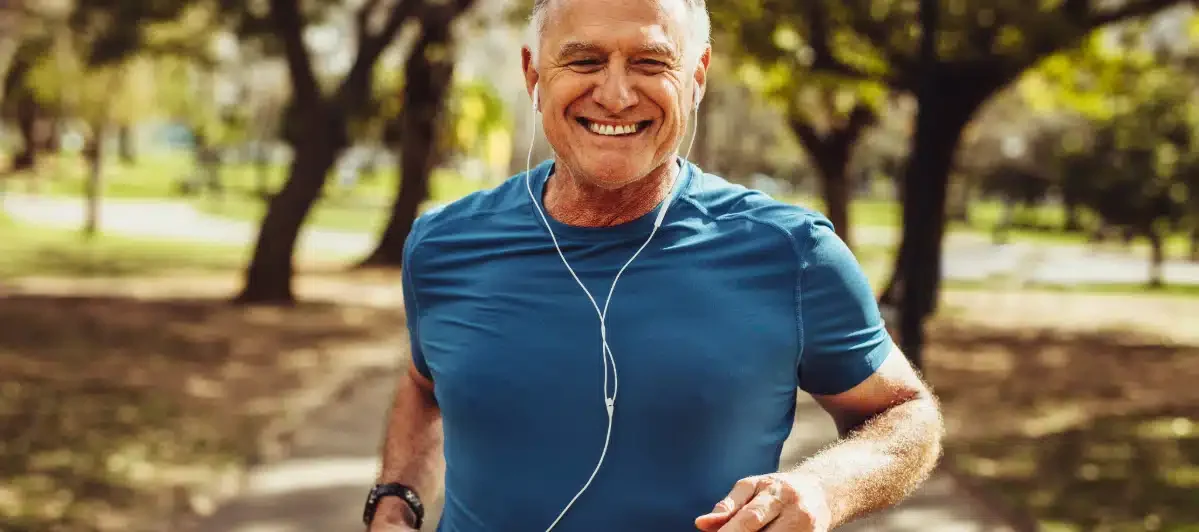 An older man jogging outdoors in a park, wearing a blue athletic shirt and listening to music with earphones, smiling.