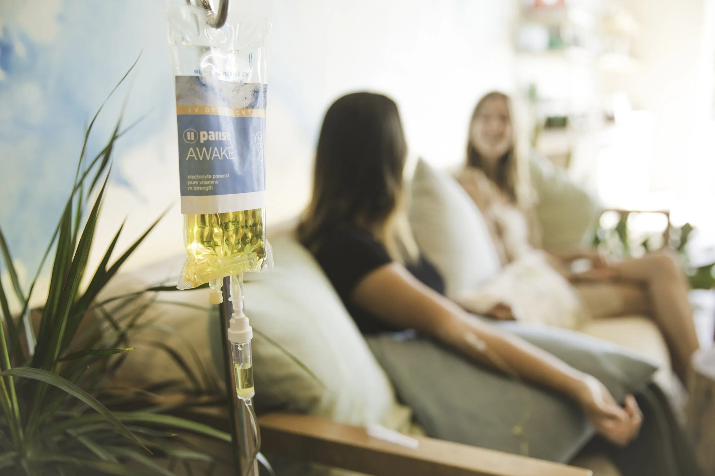 IV drip bag with yellow fluid hanging in a hospital room, with two women sitting on a couch in the background, blurred.