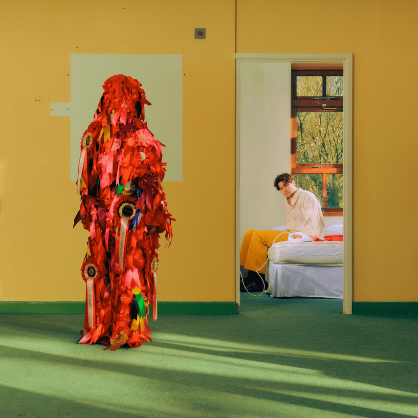 A person dressed in a colorful costume made of red and pink fabric with ribbons, standing in a room with green carpet and yellow walls. Another person sits on a bed in a nearby room visible through an open door, looking towards the costume.