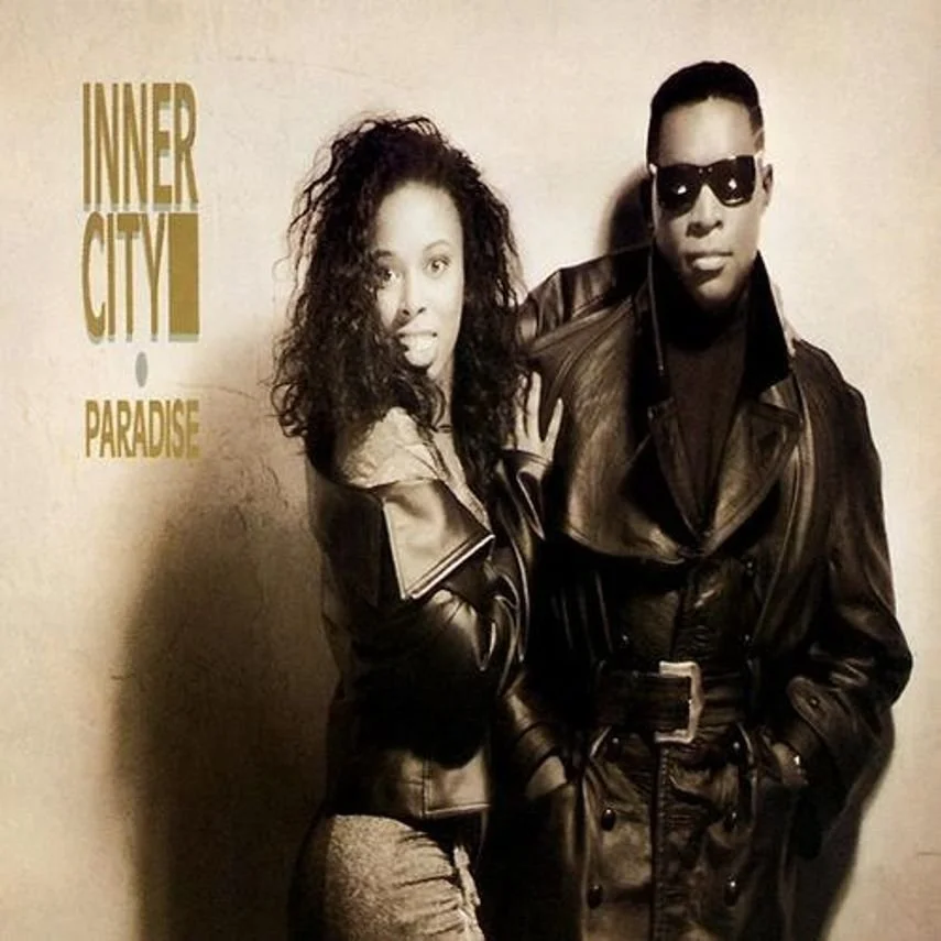 A man and woman in leather jackets, with the woman extending her arm and the man wearing sunglasses, standing in front of a wall with the words "INNER CITY PARADISE" written on it.