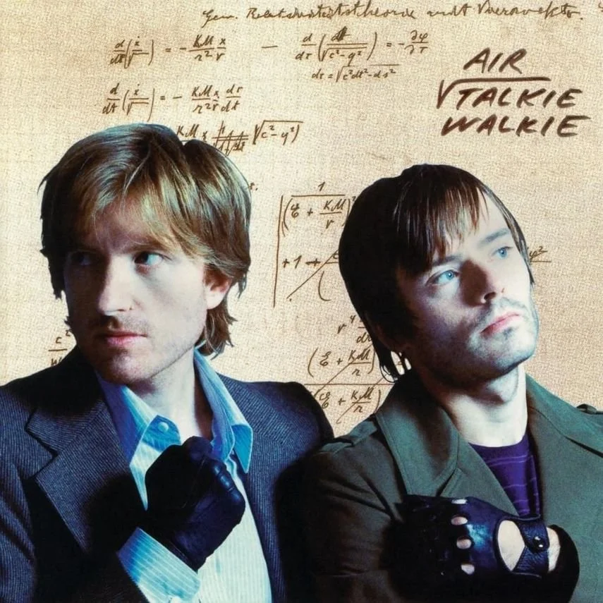 Two men with serious expressions, one with light brown hair and the other with dark hair, are dressed in formal and military-like attire respectively. Behind them, a beige background features handwritten mathematical equations and text reading 'AIR WALKIE TALKIE' in stylized font.