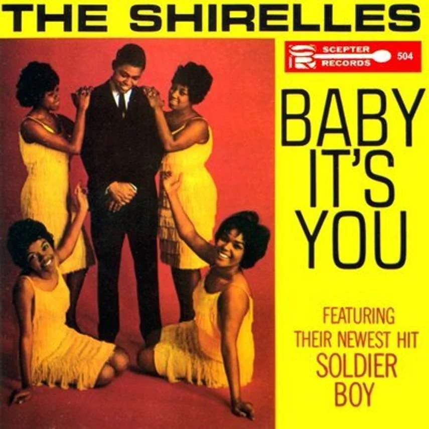 Album cover for The Shirelles' single "Baby It's You" featuring five women in yellow dresses and one man in a black suit, smiling and posing against a red background with yellow borders and black text.