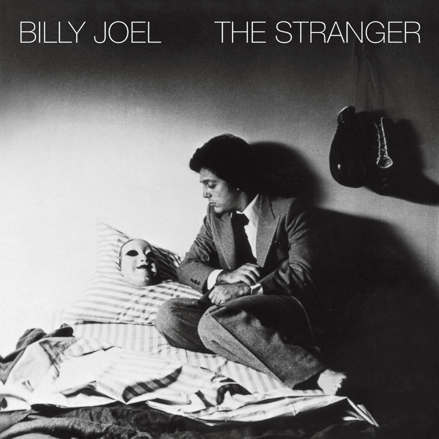 Black and white album cover of Billy Joel's 'The Stranger' featuring a person in a suit sitting on a bed, looking at a doll or mask with a smiling face wrapped in striped cloth, inside a room with a hat hanging on the wall.
