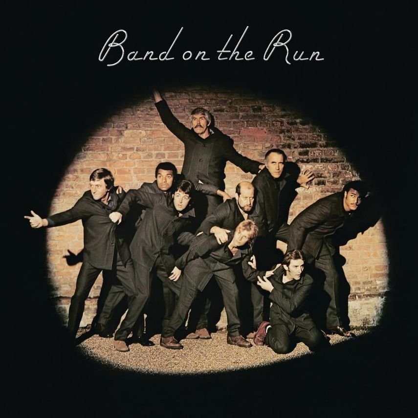 A group of people posing for a photo against a brick wall, with a spotlight creating a circle of light around them. The text 'Band on the Run' is written above.