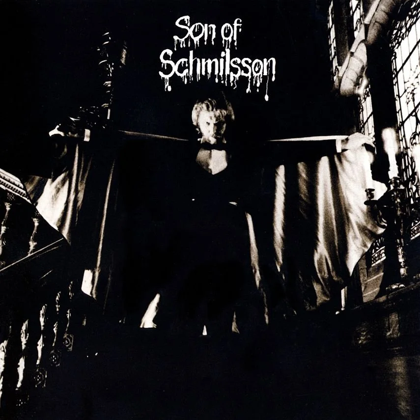 Black and white photo of a person standing on stairs, with the text 'Son of Schmilsson' written above their head, in a creepy font. The person appears to be in a dark room with ornate railings and draped curtains.