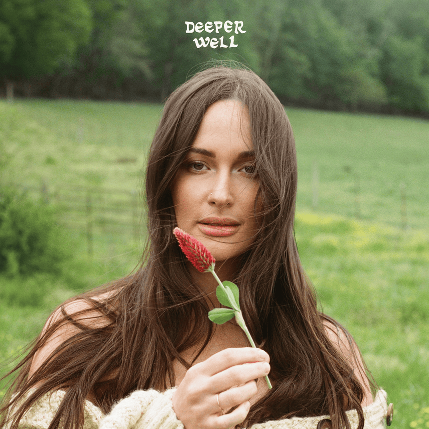 A woman with long brown hair holding a pink flower outdoors in a grassy area with trees in the background. Text says 'Deeper Well' at the top.