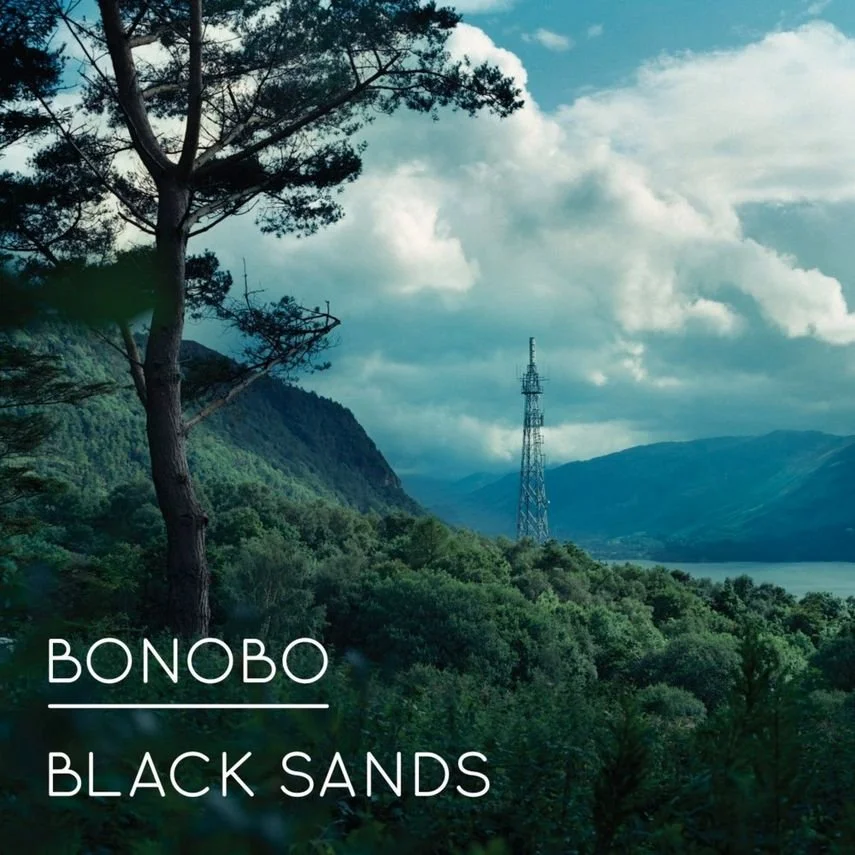 A landscape with green trees, a mountain, and a cloudy sky, featuring a tall communication tower in the distance. Text overlay reads 'BONBO BLACK SANDS'.