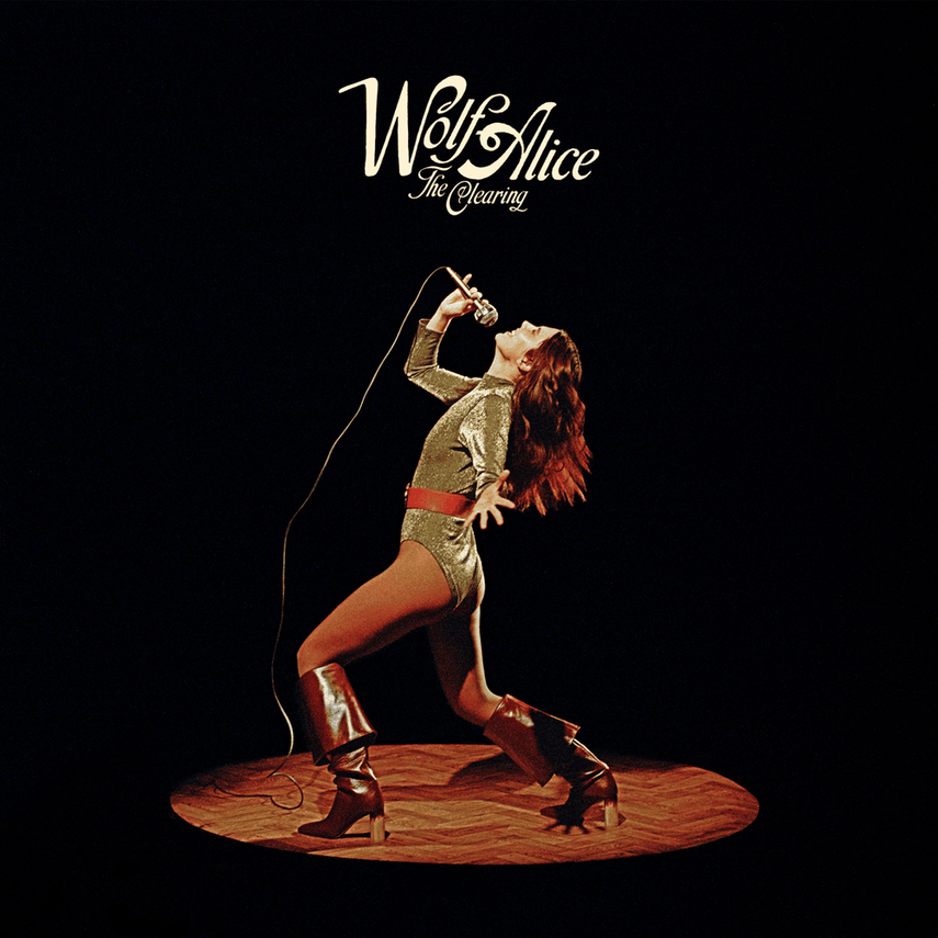 A woman with red hair in a gold outfit and boots passionately singing into a microphone on a wooden stage with a black background, with the text "Wolf Alice The Clearing" above her.