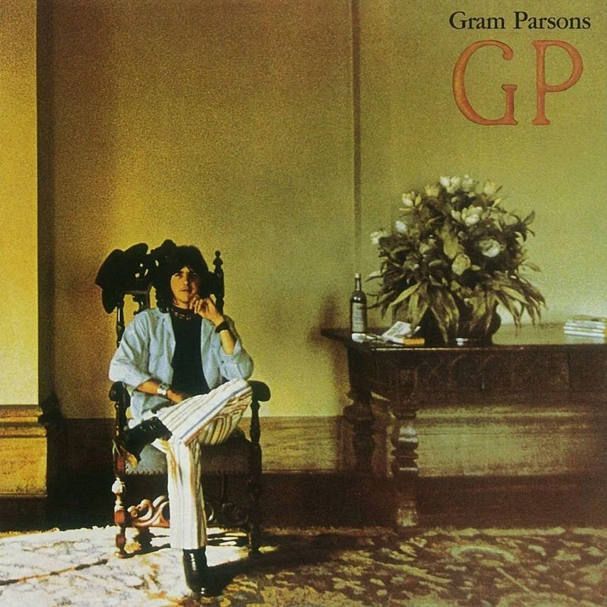 A woman sits on a wooden chair in a room with a patterned carpet, a side table with a large flower arrangement, a wine bottle, some papers, and a book. The wall behind has the name 'Gram Parsons' and the initials 'GP' in large letters.