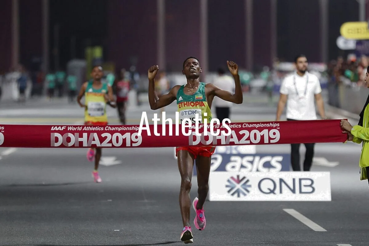 A male Ethiopian marathon runner crosses the finish line at the 2019 IAAF World Athletics Championships in Doha, tears on his face, wearing a green tank top, red shorts, and pink running shoes, with other runners and officials in the background.