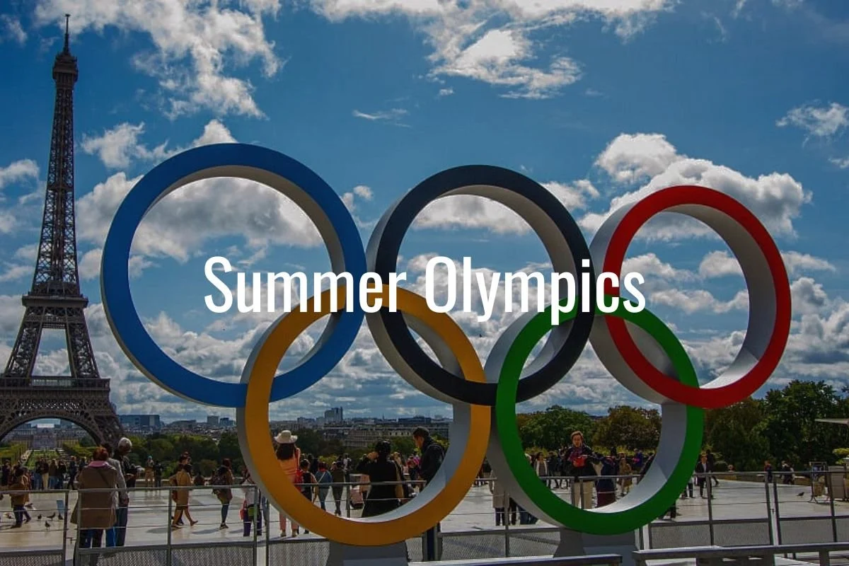 The Eiffel Tower is visible in the background under a partly cloudy sky. In the foreground, there are large Olympic rings with the words "Summer Olympics" written across them, and people are walking behind a railing.