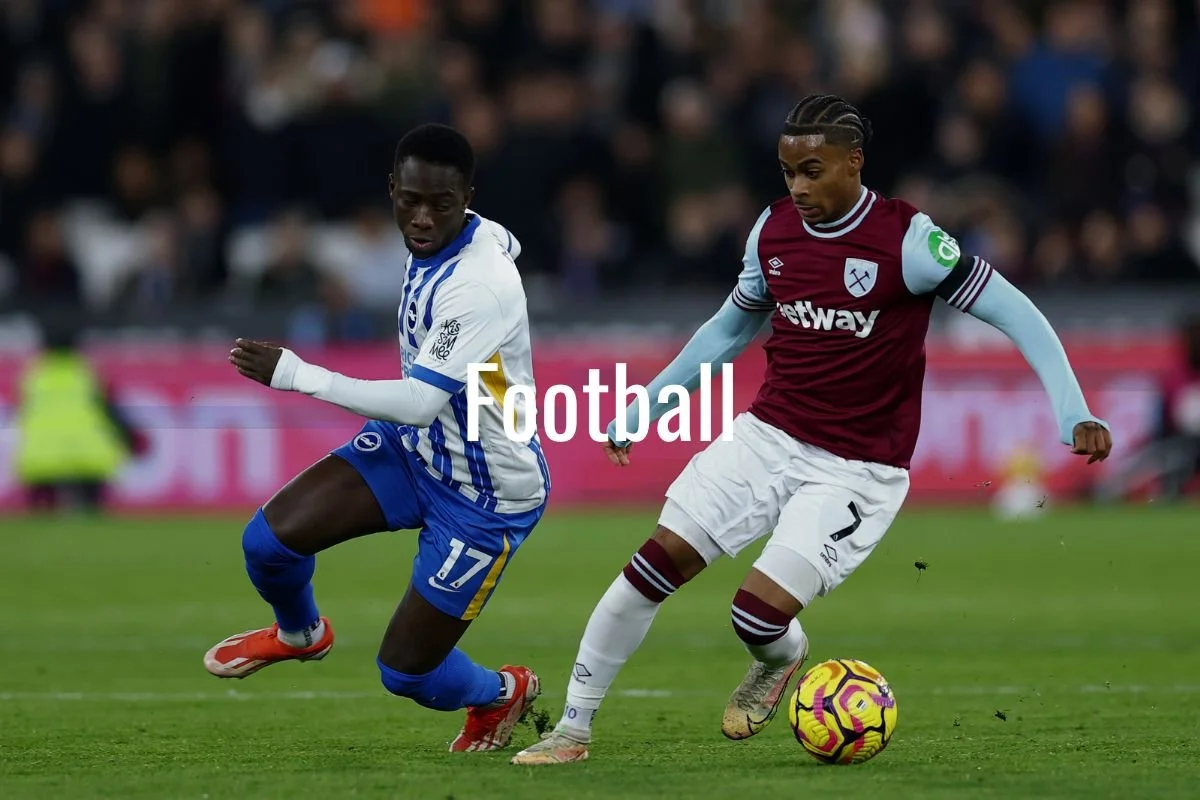 Two soccer players are competing for the ball on the field during a match. One player wears a white and blue uniform, and the other wears a claret and blue uniform. Spectators are visible in the background.