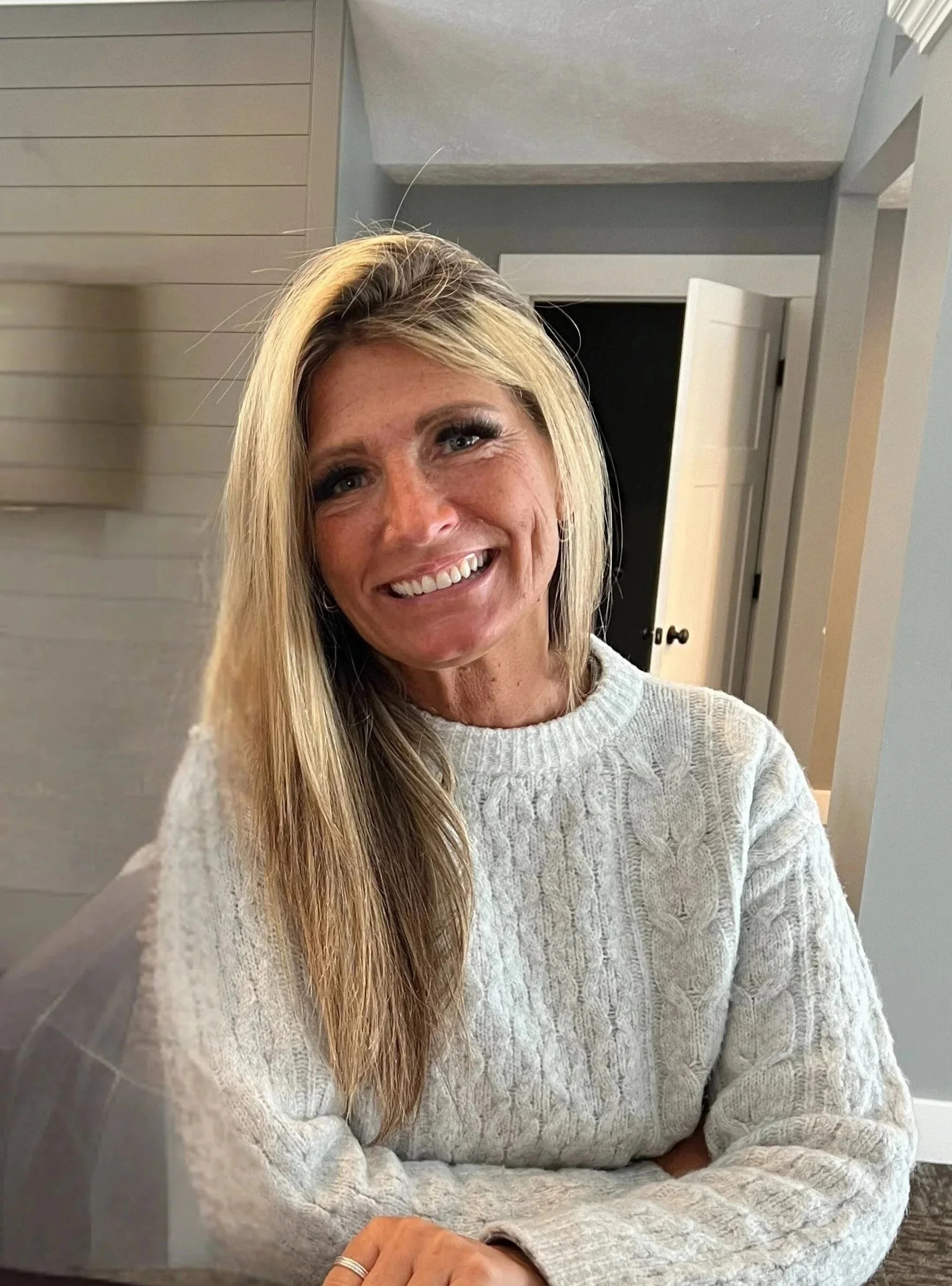 A woman with blonde hair smiling, wearing a white cable-knit sweater, sitting indoors in front of a gray and beige wall with a doorway in the background.