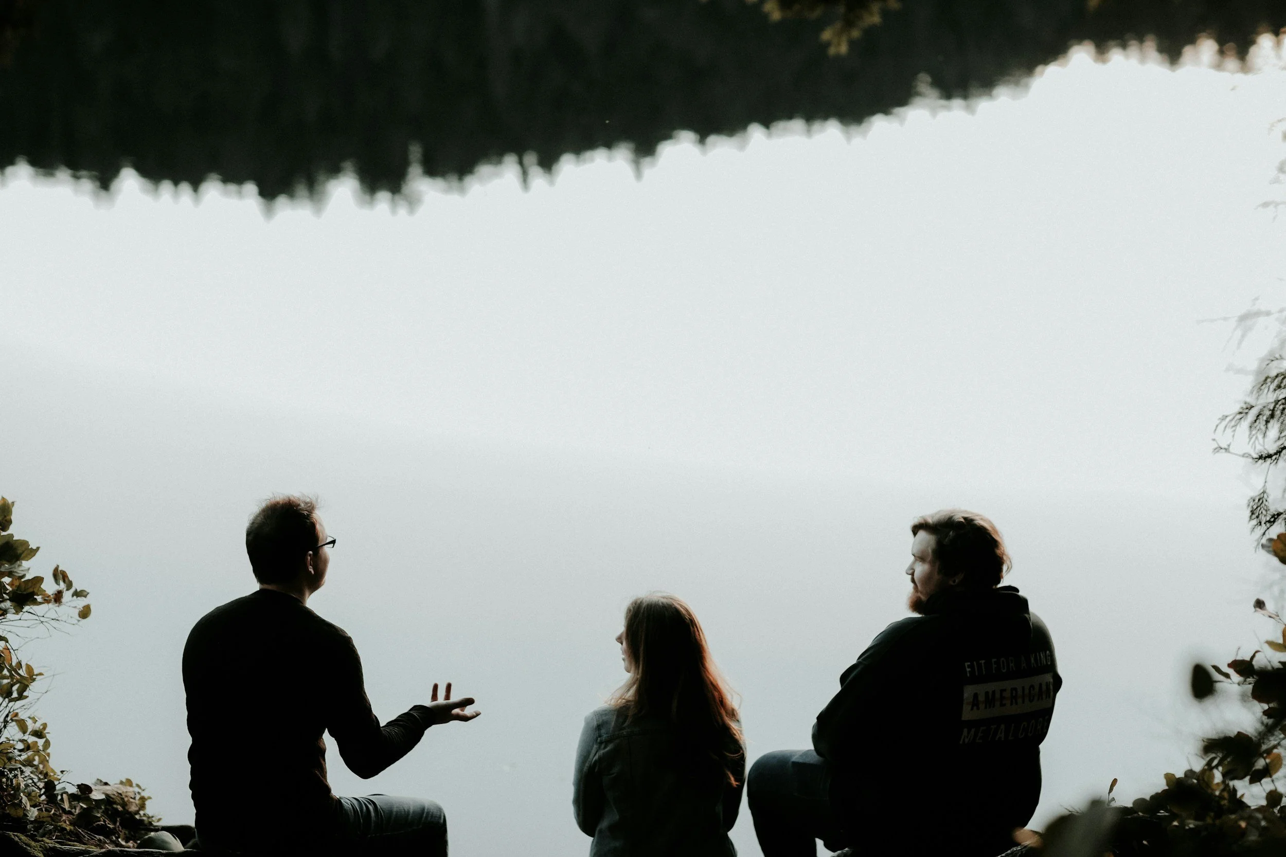 Three people sitting outdoors, engaged in conversation near water, with leaves framing the scene.