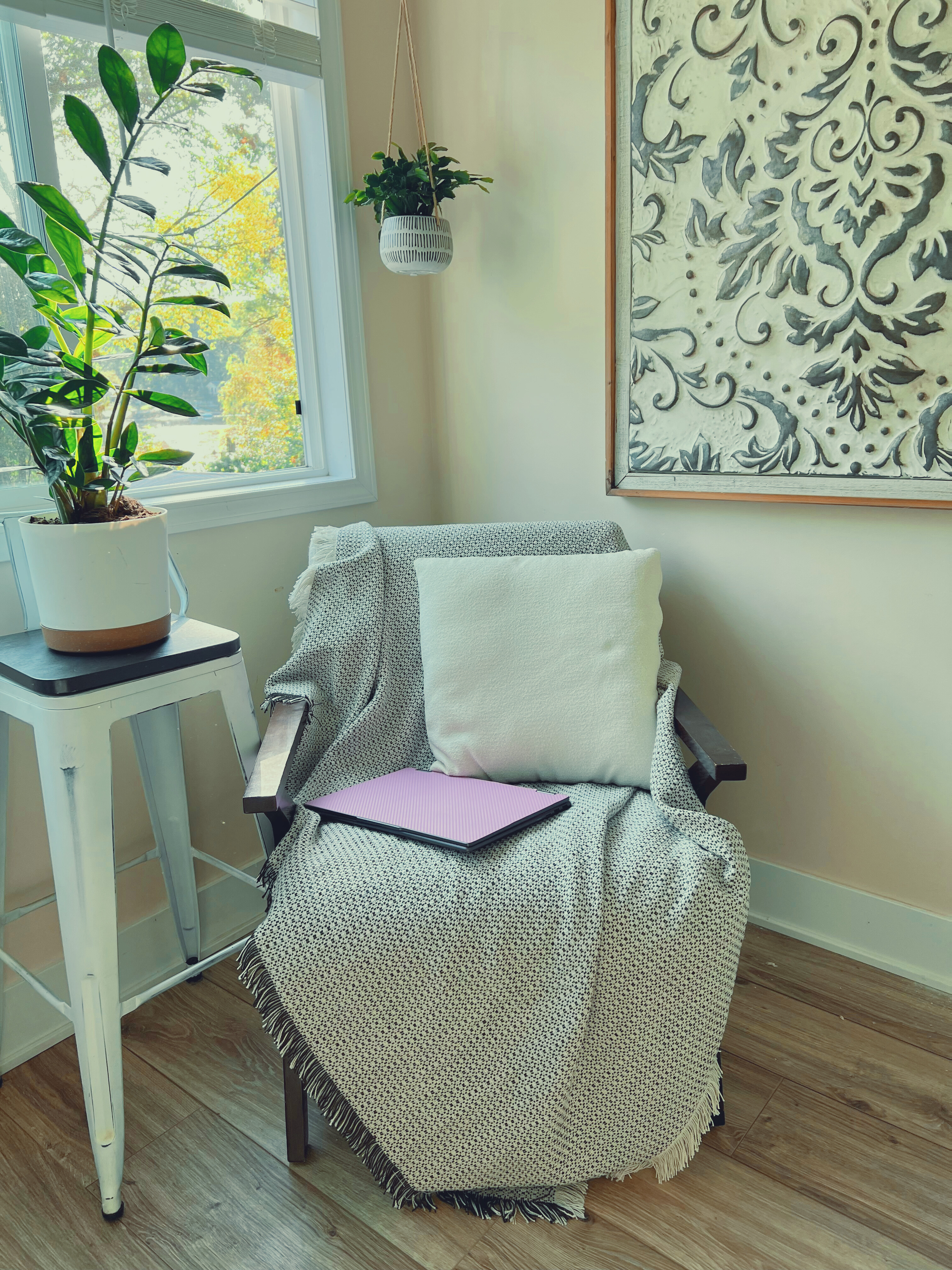 A cozy corner with a cloudy day visible outside. A window with a white frame shows trees with green and yellow leaves. Inside, a white side table holds a large potted plant with dark green leaves. Next to it, a black armchair is draped with a textured white throw blanket and has a white pillow. On the chair's seat, there is a closed purple laptop. A hanging plant in a white pot is suspended from the ceiling nearby. A large decorative wall panel with black intricate patterns on a white background is on the right side of the image.