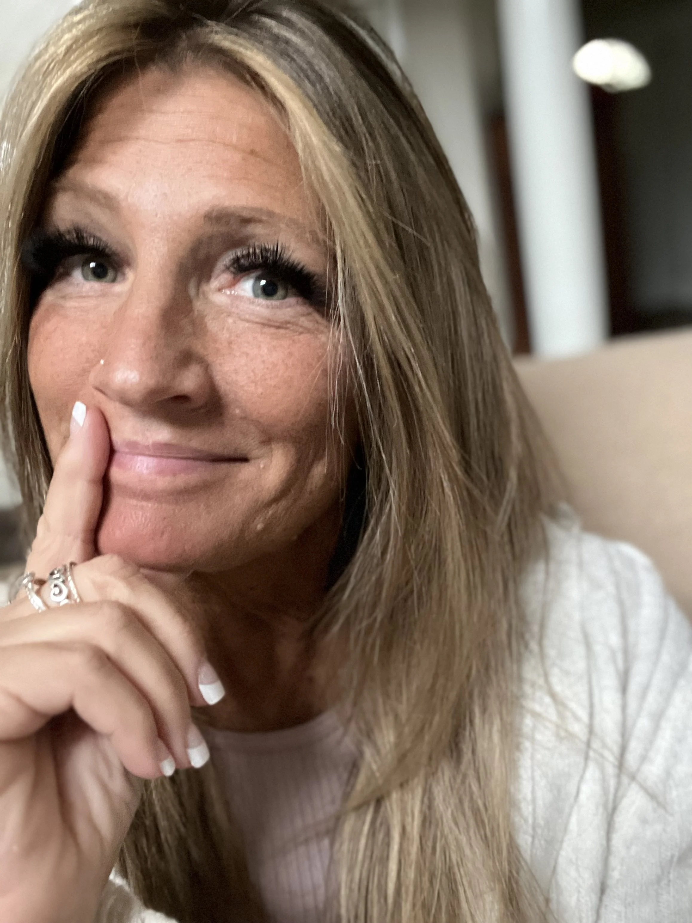 A woman with long blonde hair, blue eyes, and a nose piercing, smiling softly and touching her face with her left hand, wearing rings and a light-colored top.