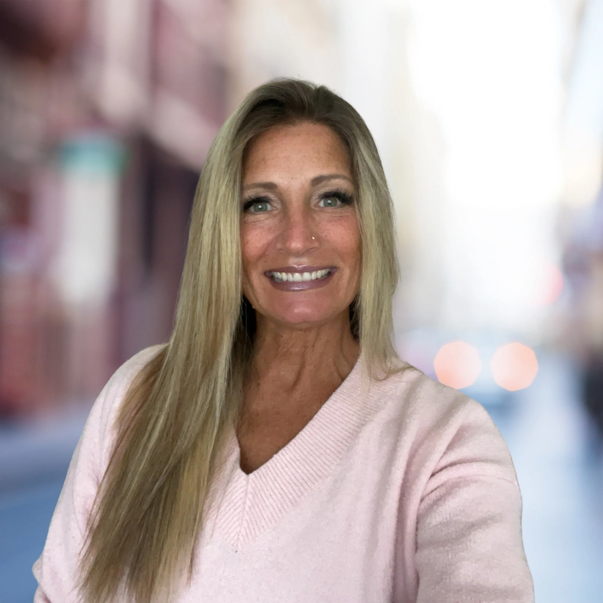 A smiling woman with long blonde hair, wearing a light pink sweater, standing outdoors with a blurred city street background.