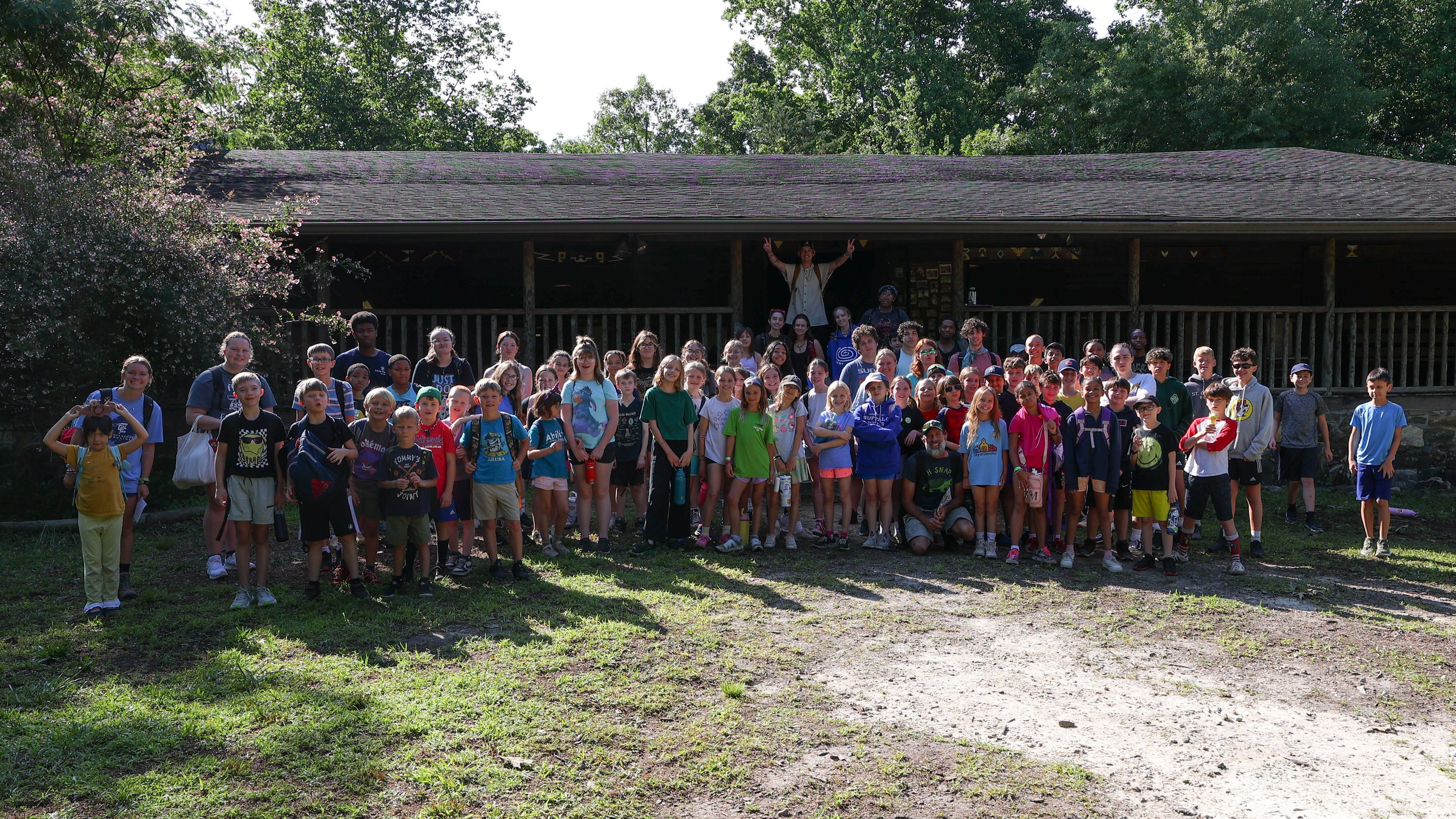 Group of children and adults gathered outdoors in front of a wooden cabin, posing for a group photo on a sunny day.