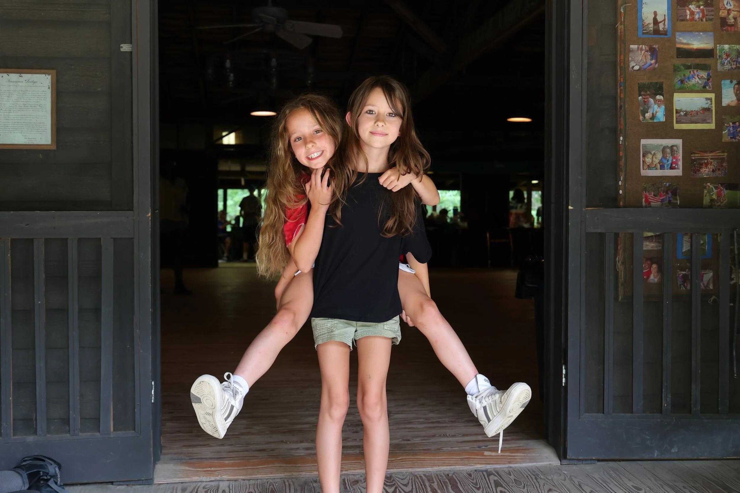 Two young girls in casual clothing, one carrying the other on her back, standing at the entrance of a rustic indoor space with photos on the wall.