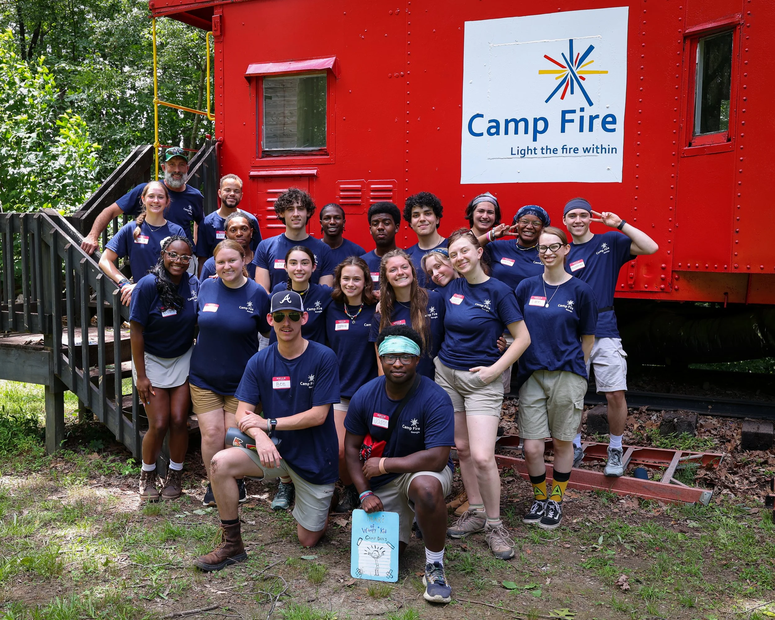 A group of young people and staff members posing outside in front of a large red train car with a Camp Fire sign. The group is smiling, some making peace signs, and everyone is wearing navy blue T-shirts with the Camp Fire logo. The setting is outdoors with trees in the background.