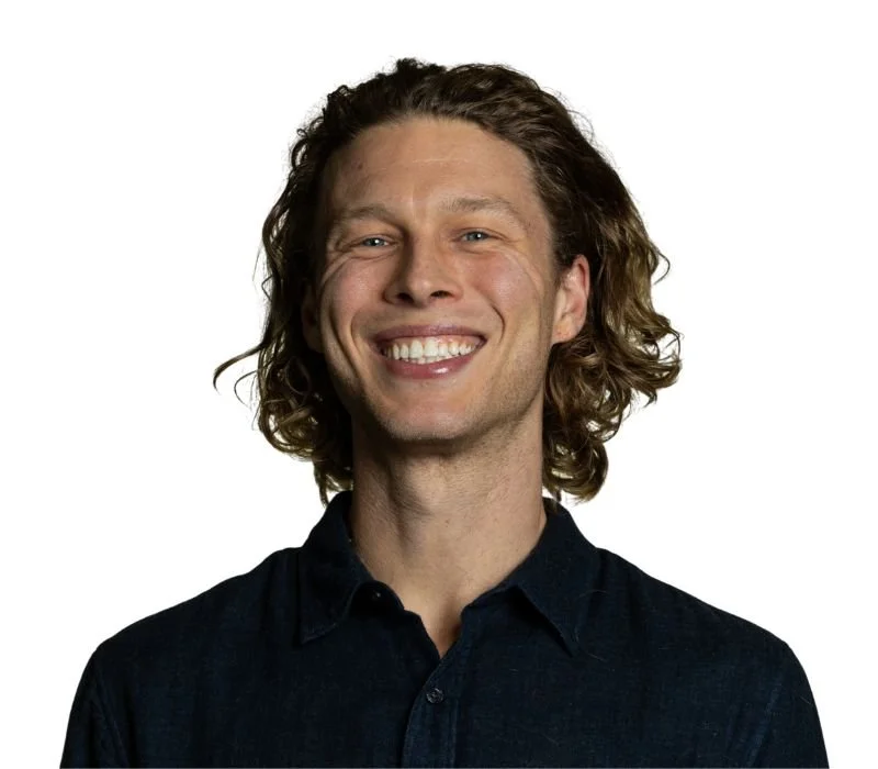 A man with long, curly brown hair smiling and wearing a dark shirt against a white background.