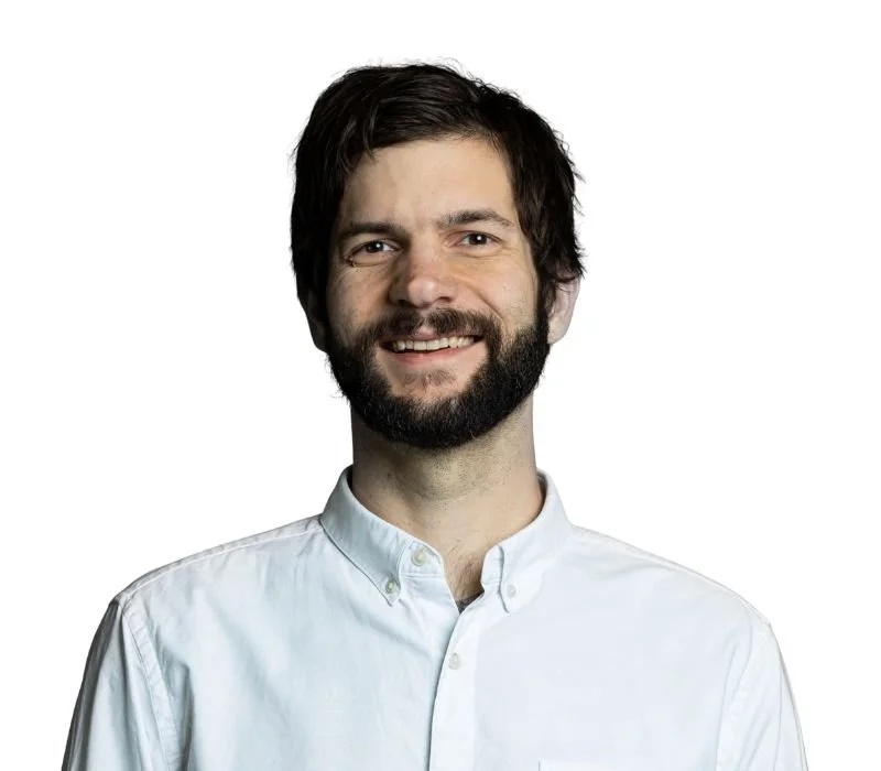 A smiling man with dark hair and beard wearing a white button-up shirt against a plain white background.