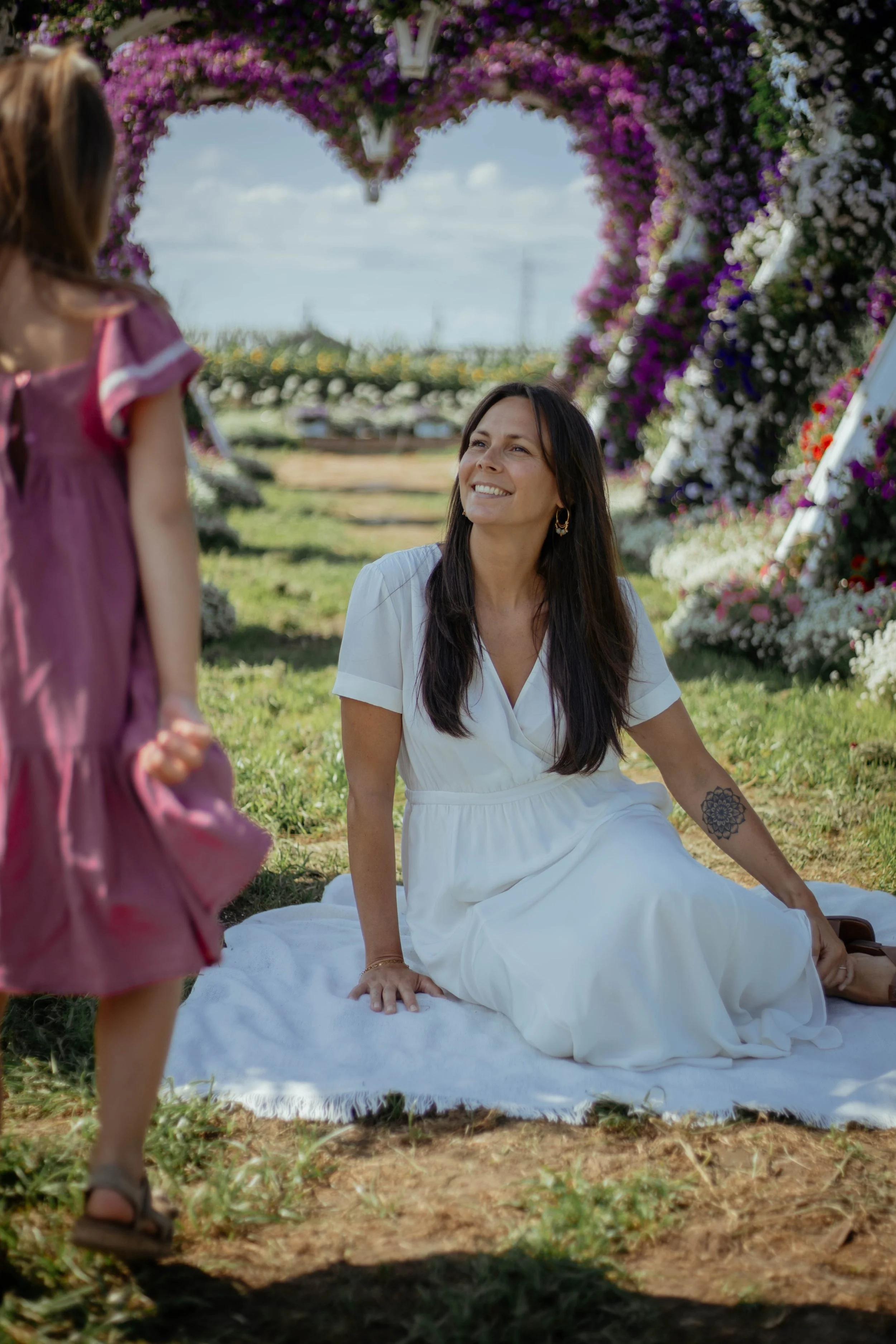 Une femme portant une robe blanche assise sur une couverture blanche dans un champ, souriant en regardant une petite fille en robe rose. Un décor floral avec un arc de fleurs violettes en arrière-plan.
