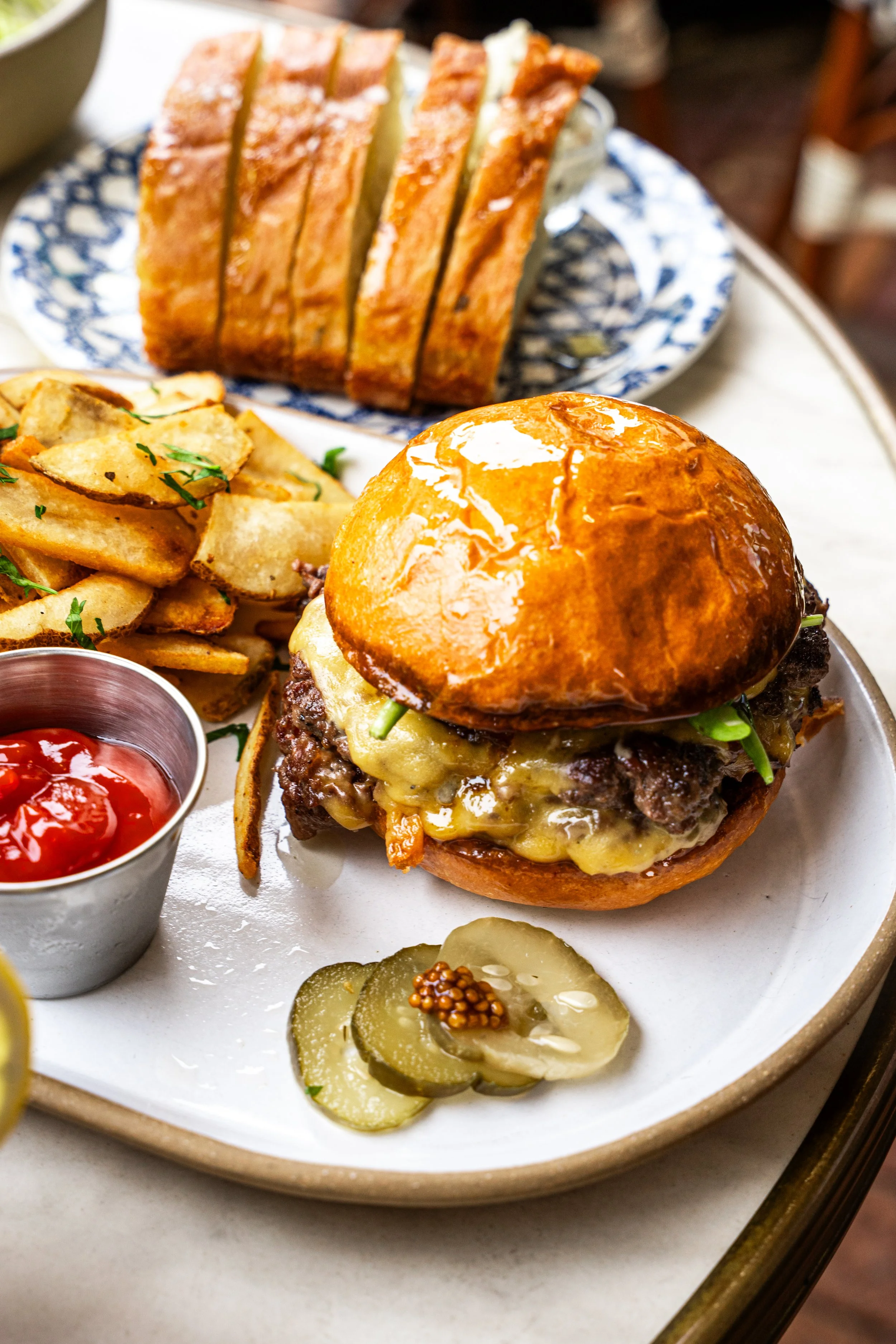 A cheeseburger with pickles, potato chips, ketchup, toasted bread, and slices of bread on a wooden table.