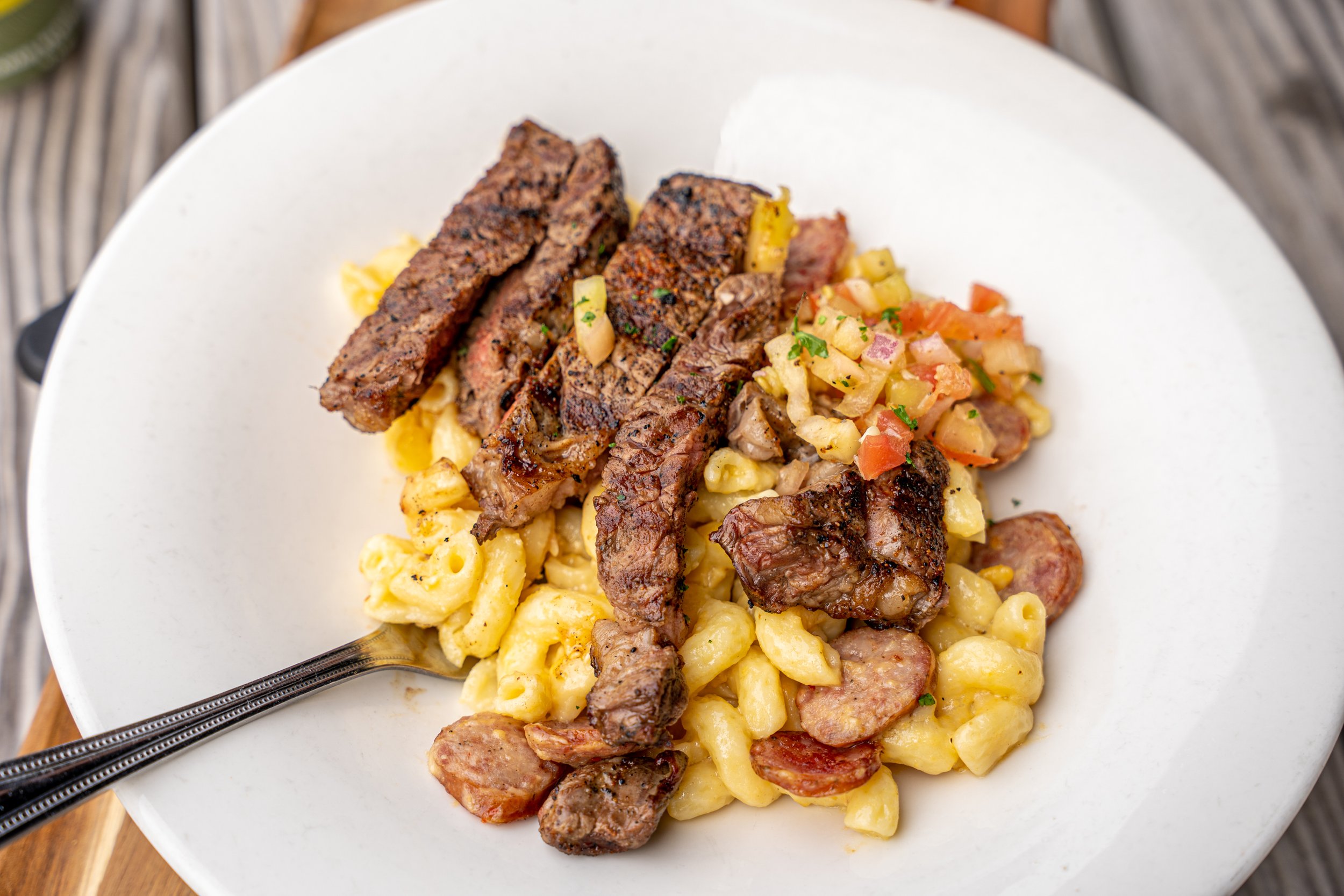 Steak slices on a bed of macaroni and cheese with sausage and vegetable salad on a white plate.