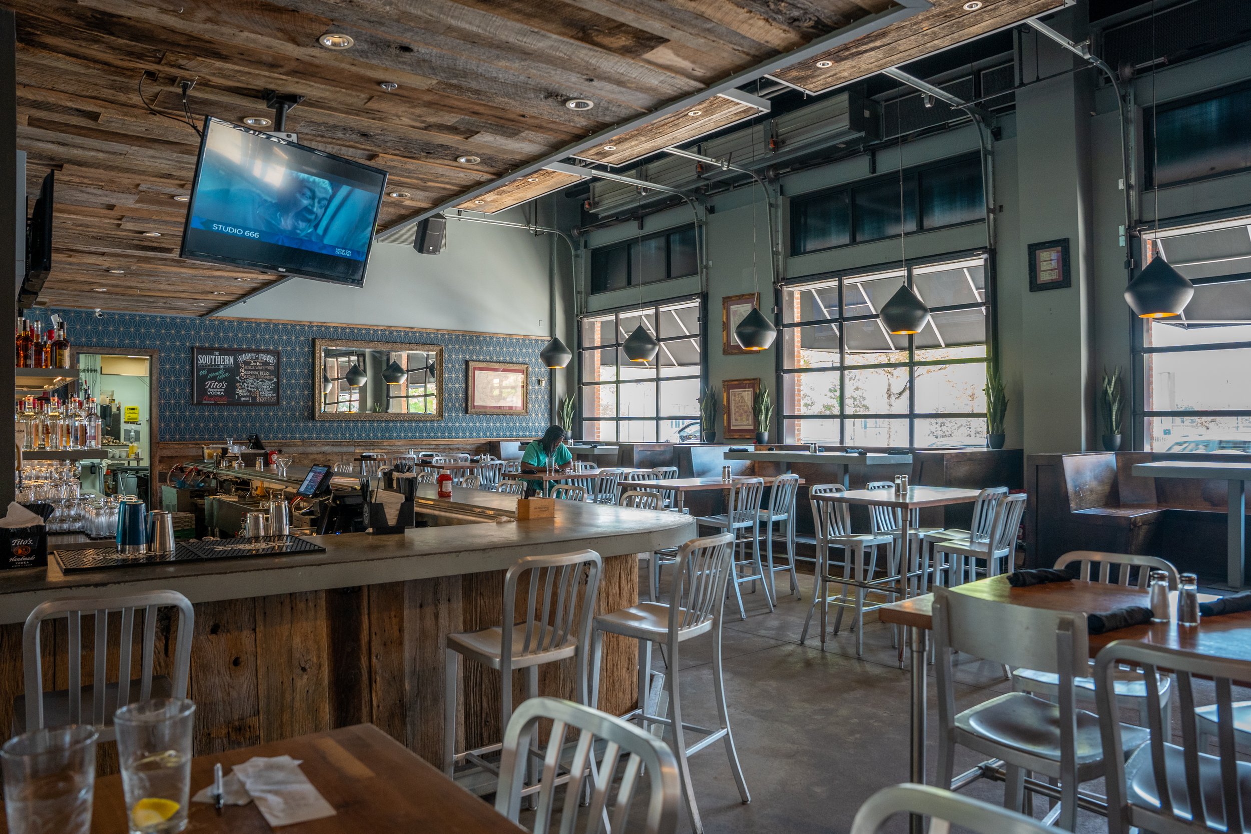Empty restaurant interior with high windows, wooden ceiling, bar counter, chairs, tables, and a person sitting at one table.
