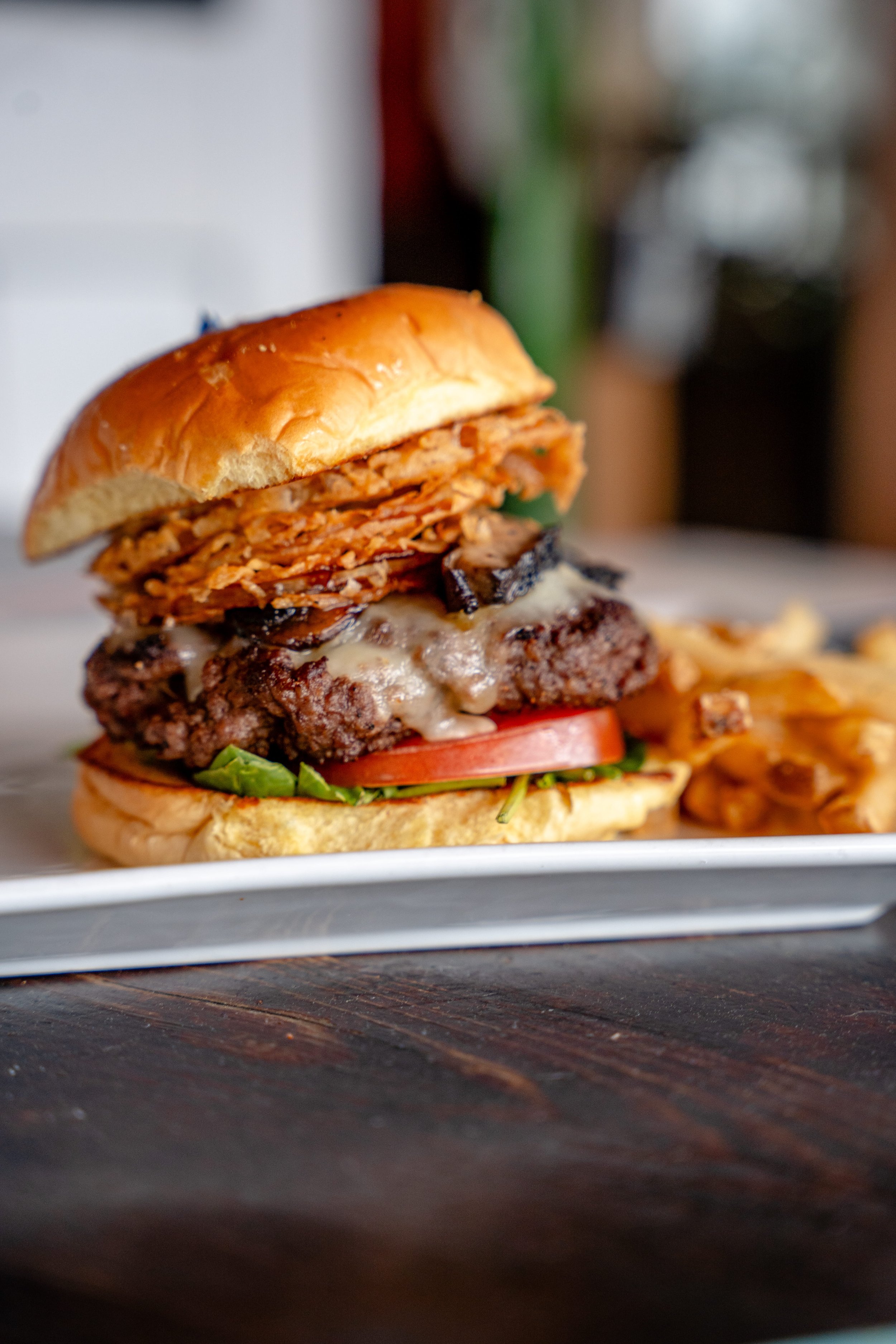 Close-up of a gourmet hamburger with a beef patty, melted cheese, grilled mushrooms, sliced tomato, lettuce, on a sandwich bun, served on a white plate with French fries in the background.