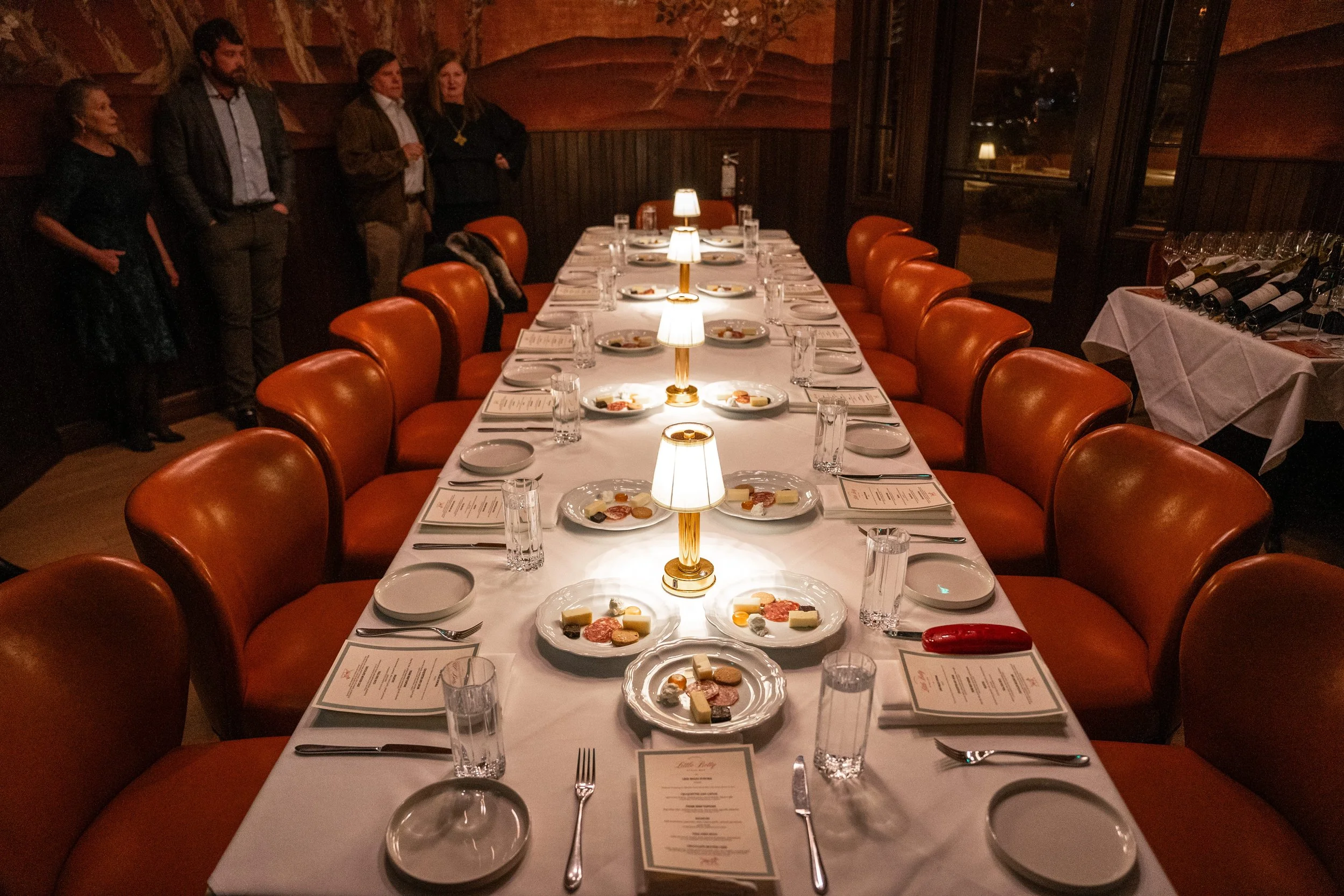 A long dining table set for a formal dinner with white tablecloths, illuminated by small lamps, surrounded by orange chairs, with plates, glasses, utensils, and menus arranged at each seat. In the background, four people are standing in a dimly lit r