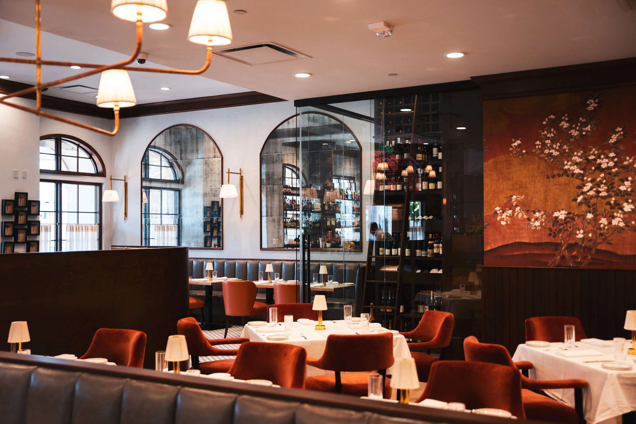 Elegant restaurant interior with white walls, large arched windows, and warm lighting. Dining tables are set with white tablecloths, glasses, and small lamps. Red velvet chairs surround the tables, and there is a large decorative painting of a flower