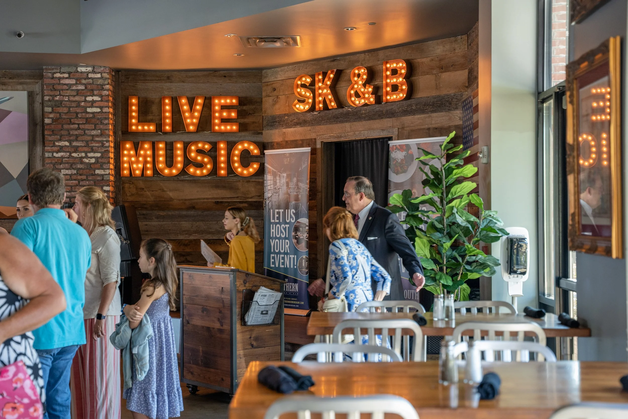 People standing in line at a restaurant or event venue with a sign reading 'LIVE MUSIC,' and a decorative wall with illuminated letters 'SK & B.' Inside, there are tables, chairs, a large green plant, and a banner that says 'LET US HOST YOUR EVENT!'