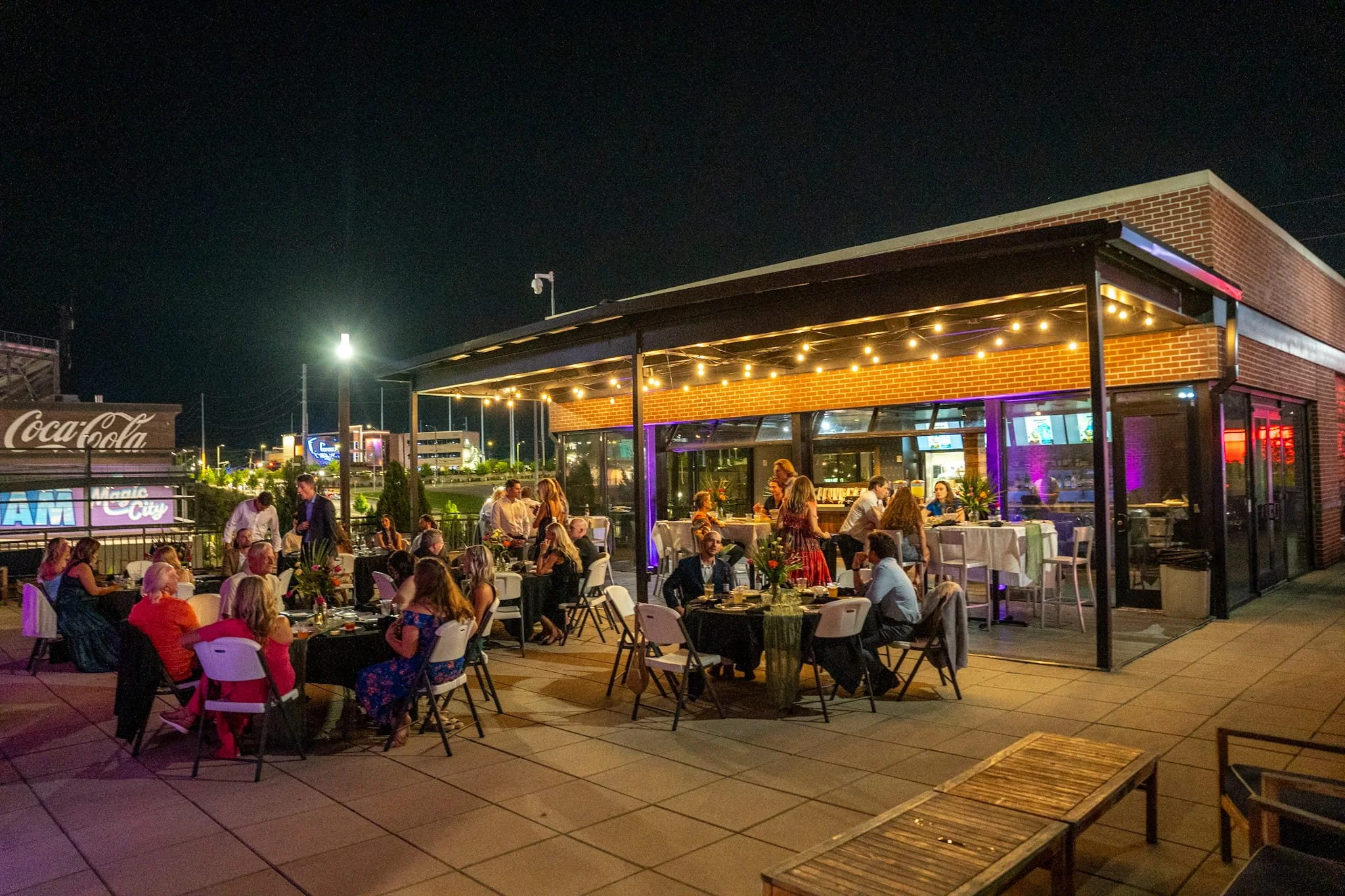 Outdoor restaurant patio at night with guests dining and socializing, illuminated by string lights with a building featuring glass walls and neon purple lights.