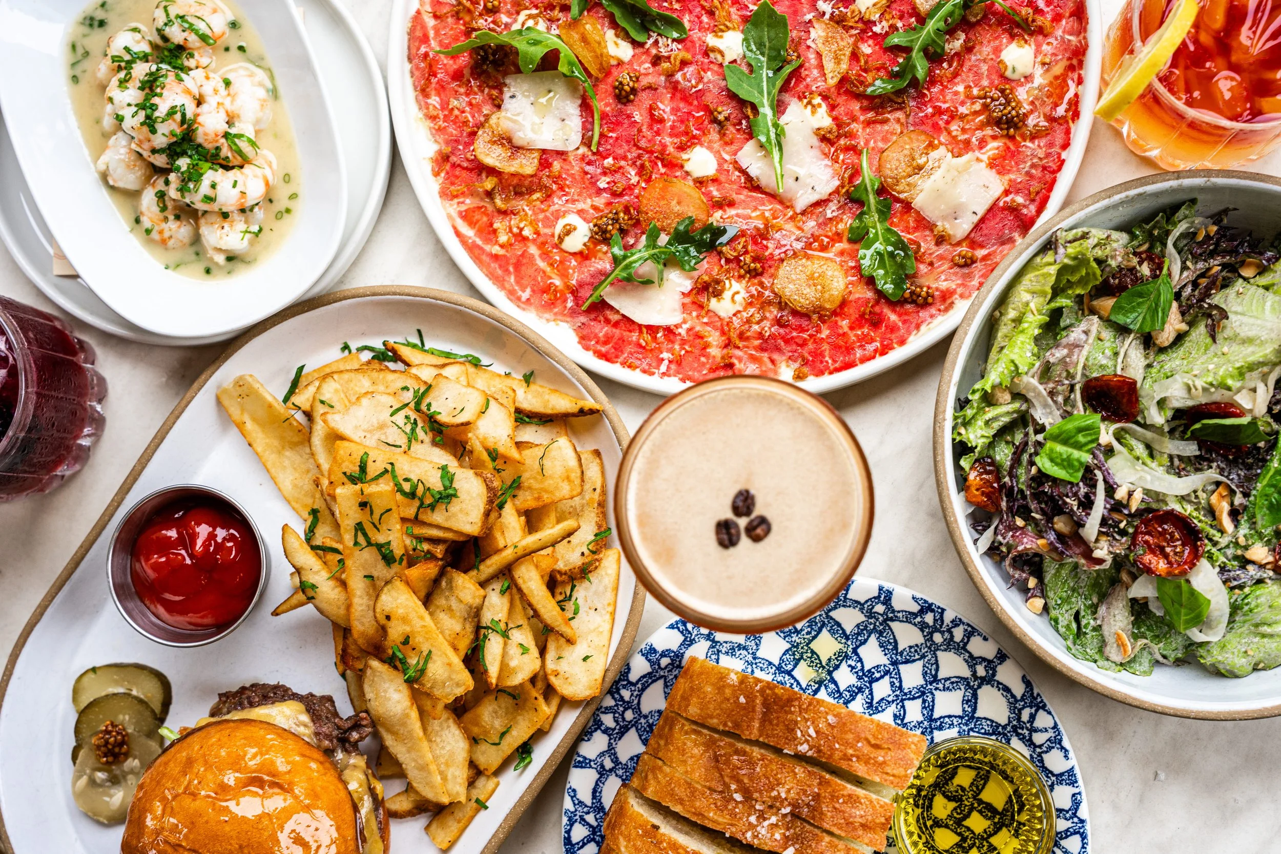 A table with various dishes including a pizza with arugula, a salad with cherry tomatoes and nuts, a bowl of shrimp, a plate of French fries, a burger, slices of bread with olive oil, and drinks like coffee and iced tea.