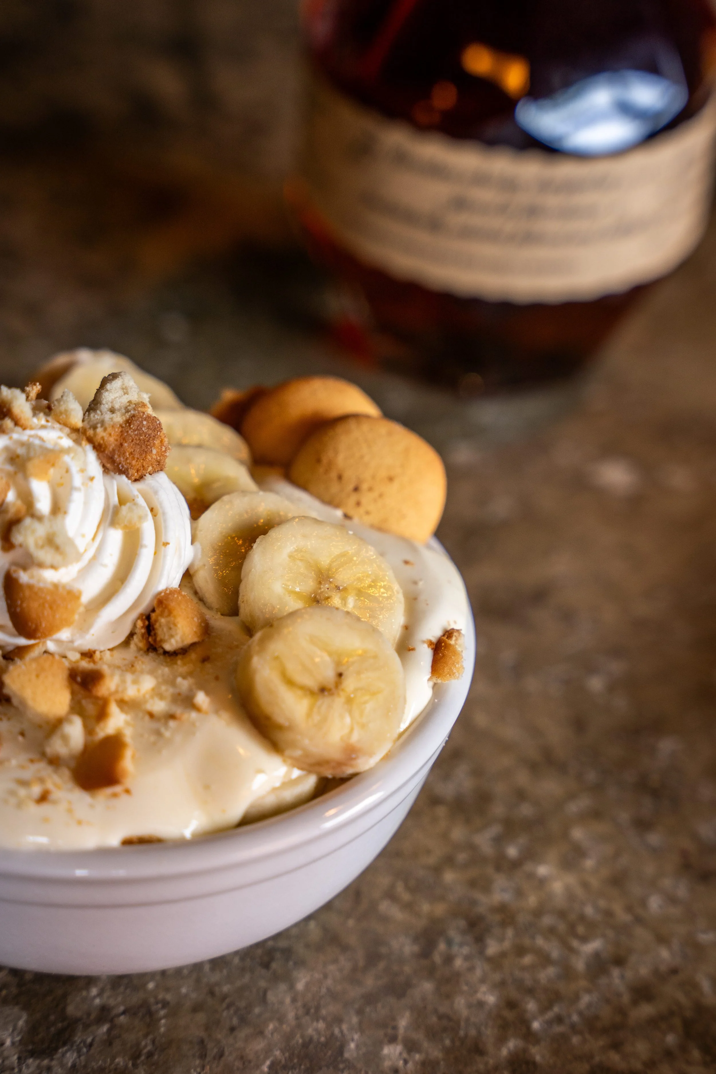 A bowl of banana pudding topped with sliced bananas, whipped cream, and crushed cookies on a marble surface with a jar in the background.