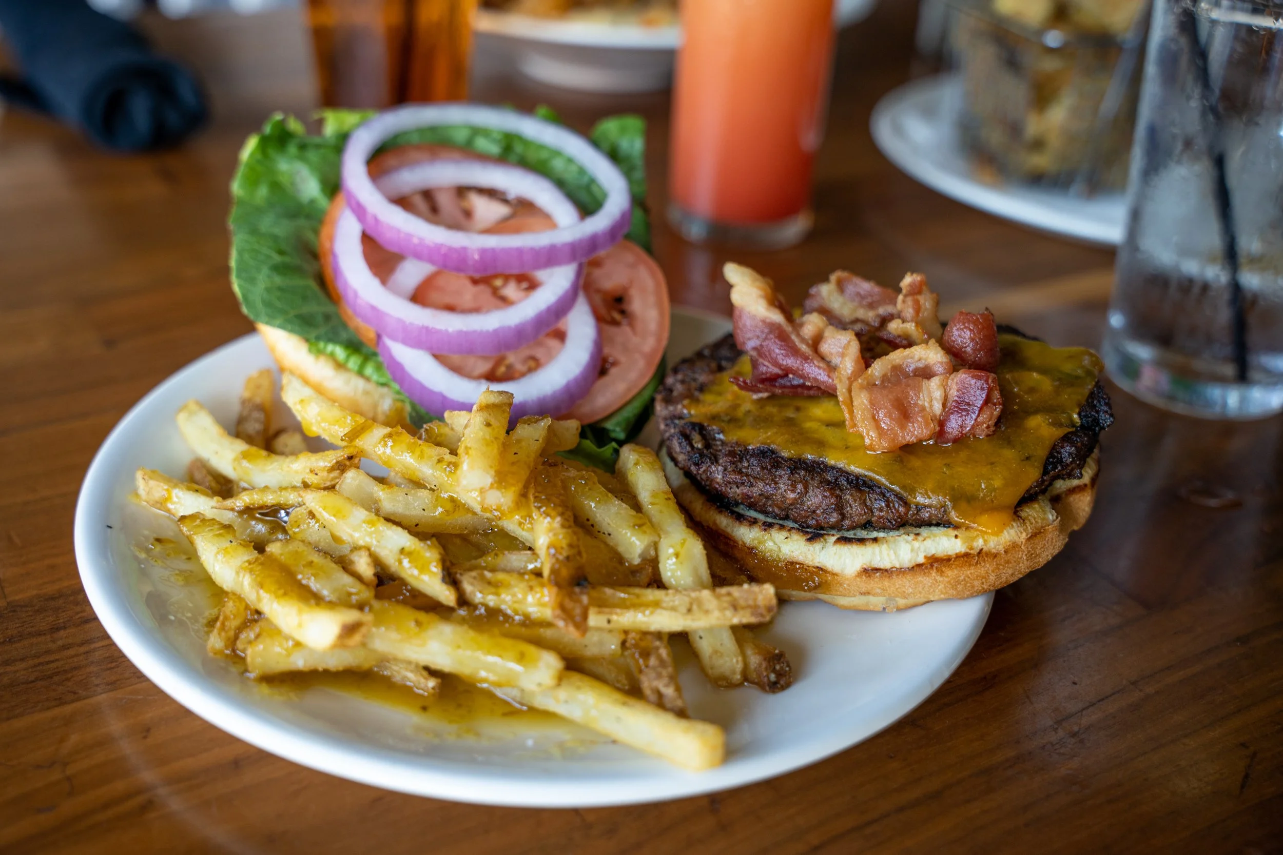 Burger with bacon, cheddar cheese, lettuce, tomato, onion, side of French fries, and a glass of iced tea on a wooden table.