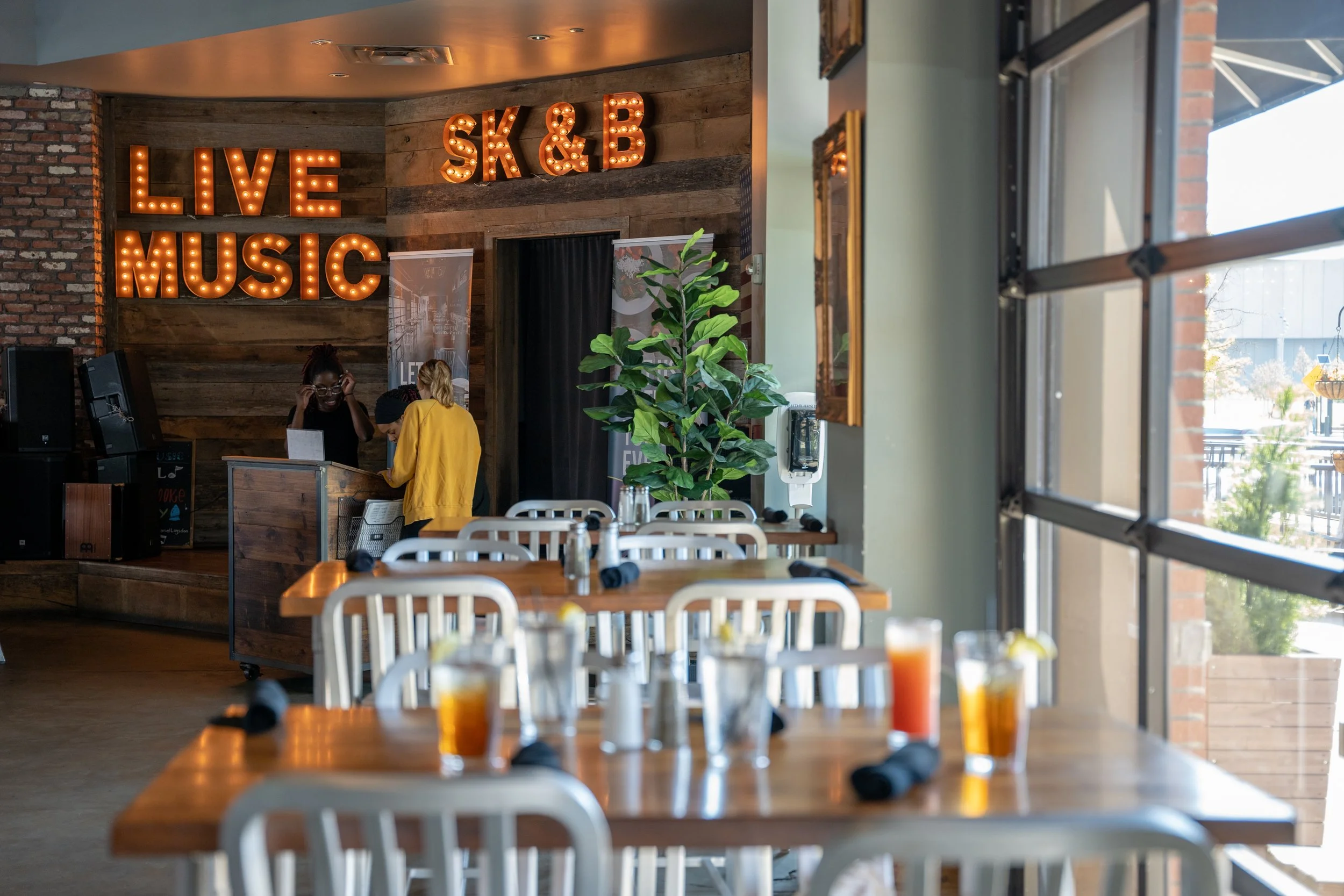 Empty restaurant with tables set with drinks and utensils, large window with sunlight, and a music stage with illuminated sign reading 'LIVE MUSIC' and 'SK & B', two people at the counter