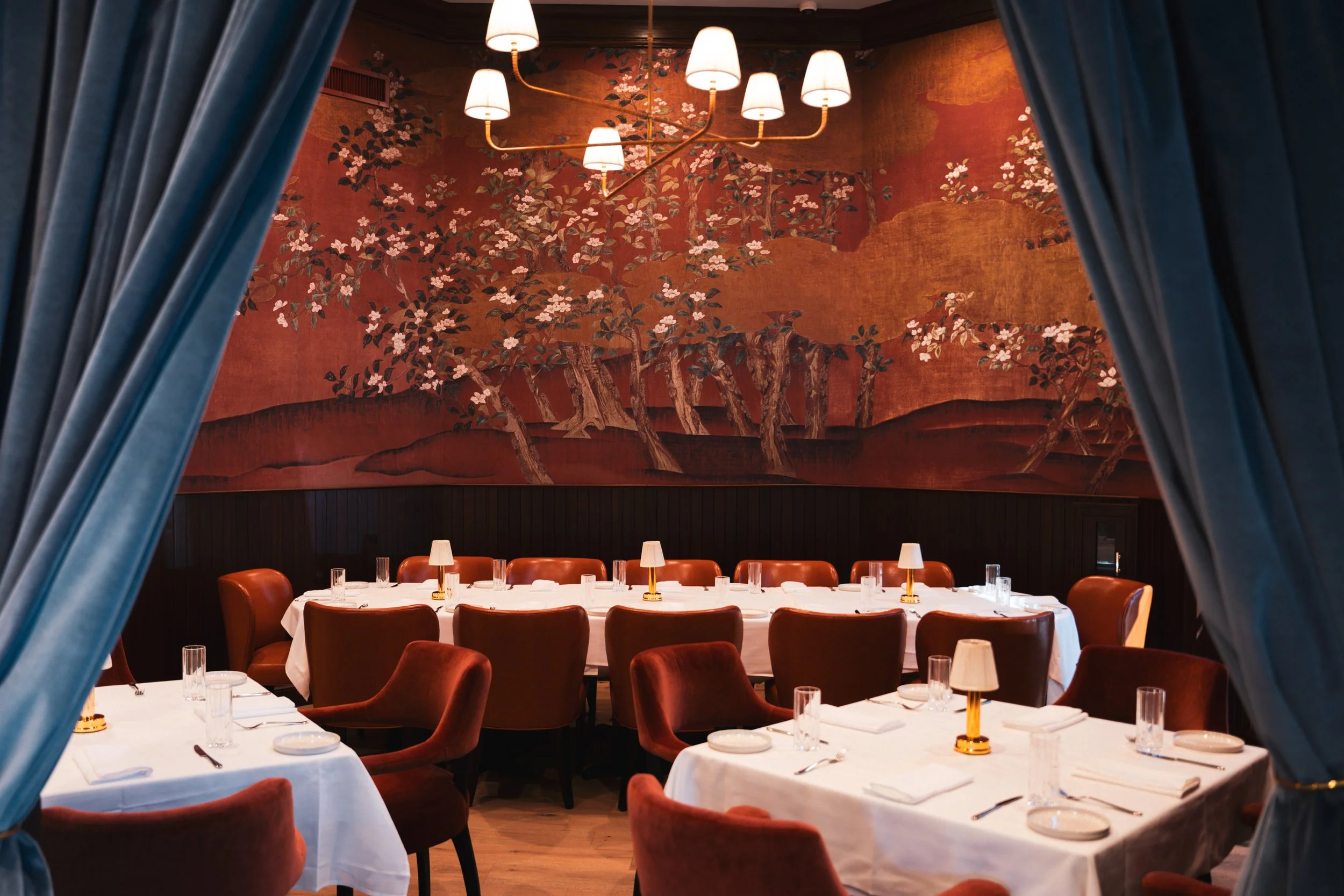 A private dining room seen through blue curtains, featuring a large oval table with white tablecloths, chairs with reddish-brown upholstery, and small lamps on the table. The wall has a large mural depicting trees and flowering branches in warm tones