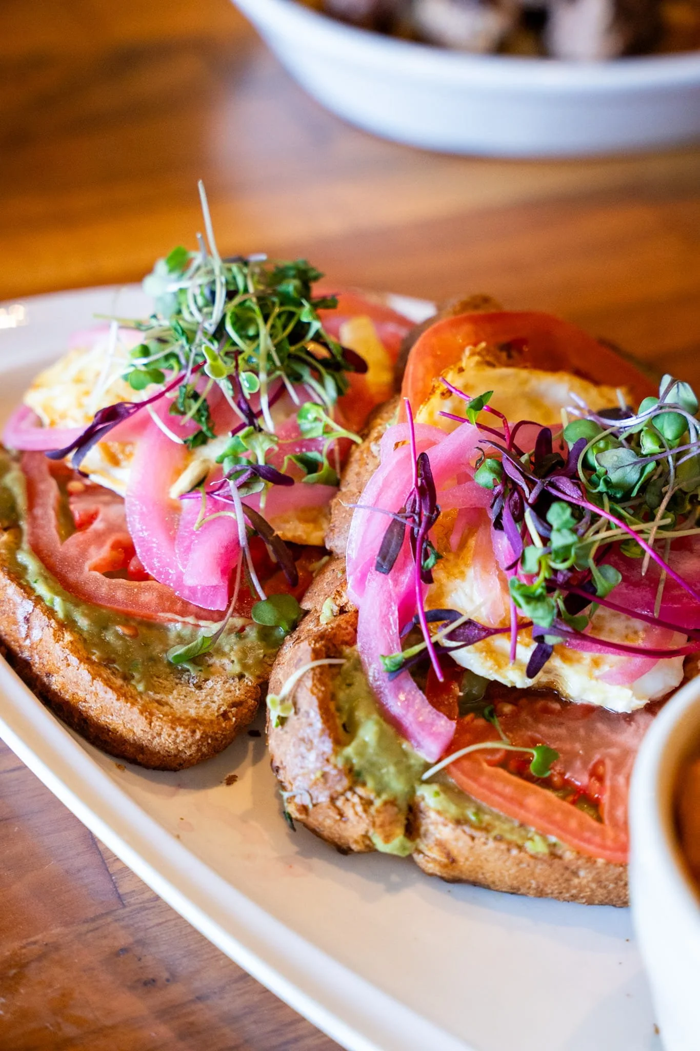 Open-faced avocado toast topped with tomato slices, pickled onions, microgreens, and herbs on toasted bread with a side of sauce.
