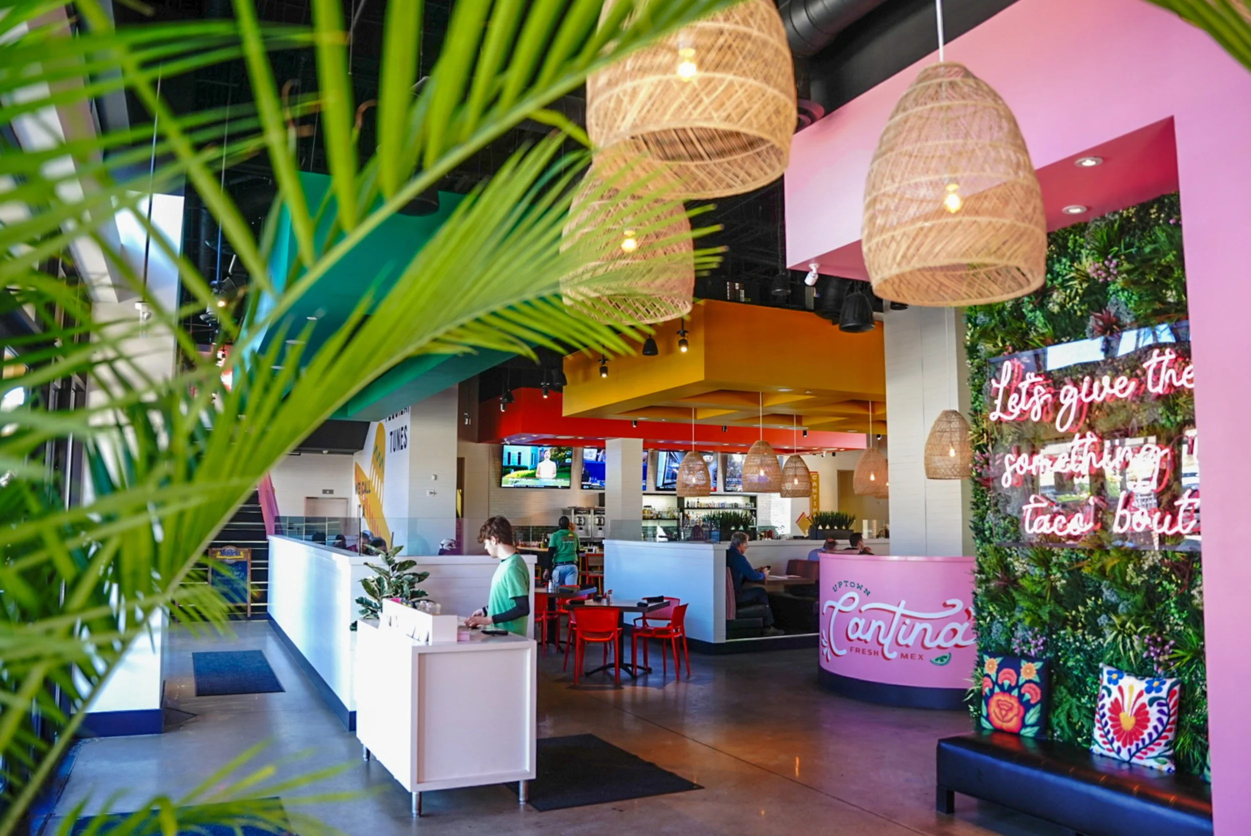 Colorful interior of a modern restaurant or cafe with hanging woven lamps, a pink curved counter with the sign 'Cantina,' and a green plant in the foreground. Features a seating area with red chairs, a bar with TVs, and neon signage.