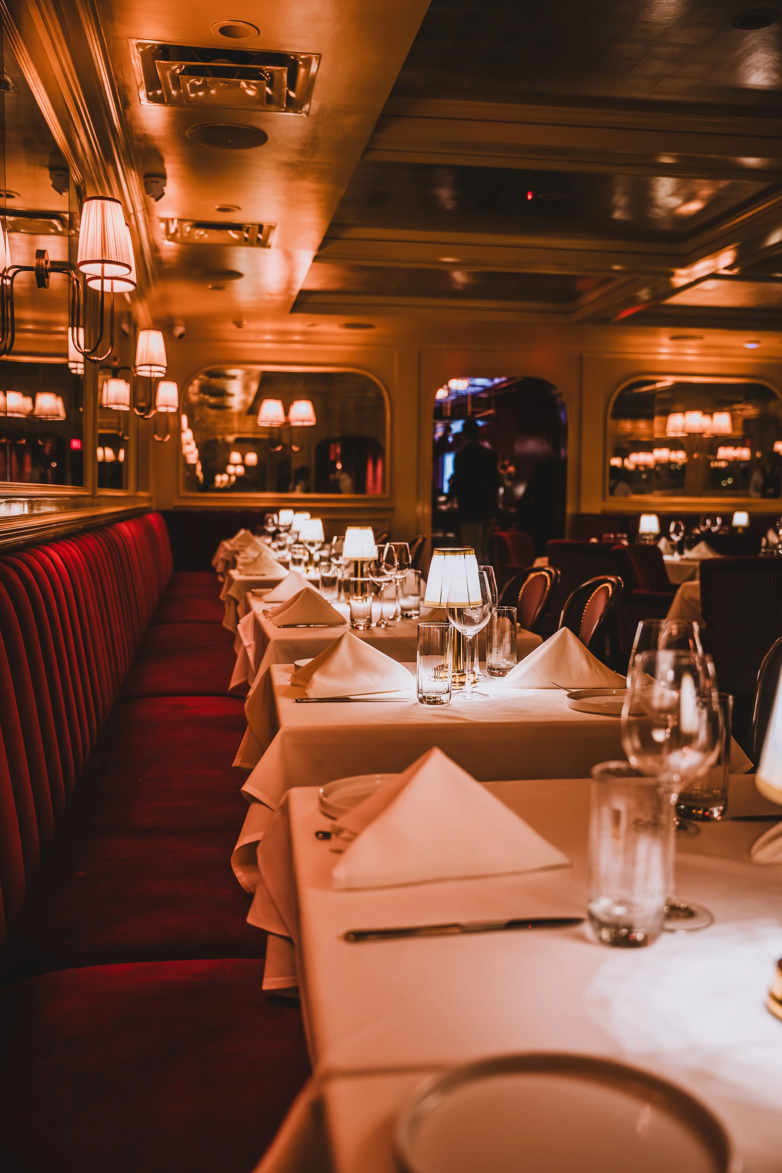 Elegantly set dining tables in a dimly lit restaurant with white tablecloths, napkins, glasses, and candles.